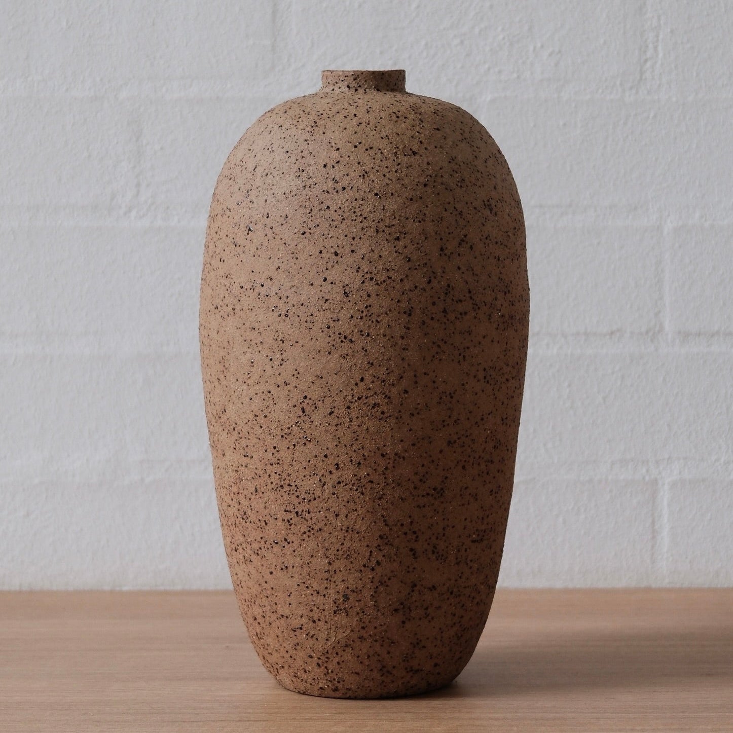 Brown speckled ceramic vessel on a wooden surface with a white textured wall background