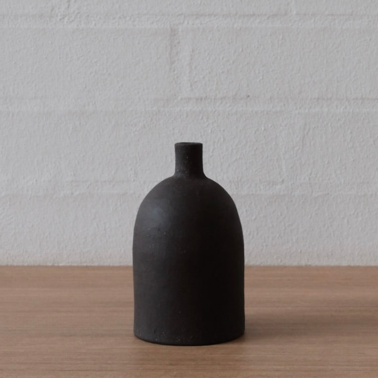 A matte black ceramic bottle with a rounded body and short neck, standing on a light wood surface against a white brick wall background.