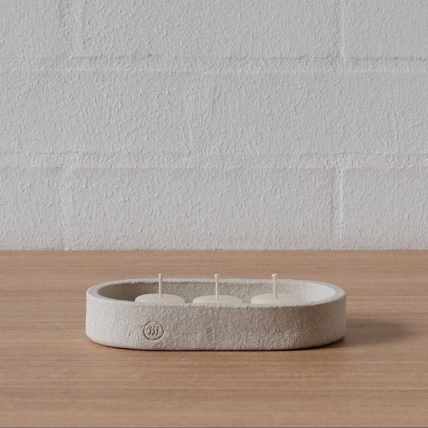 A minimalist, low-profile oval tray or shallow dish with three tea light candles inside, set on a light wood surface against a white brick wall.