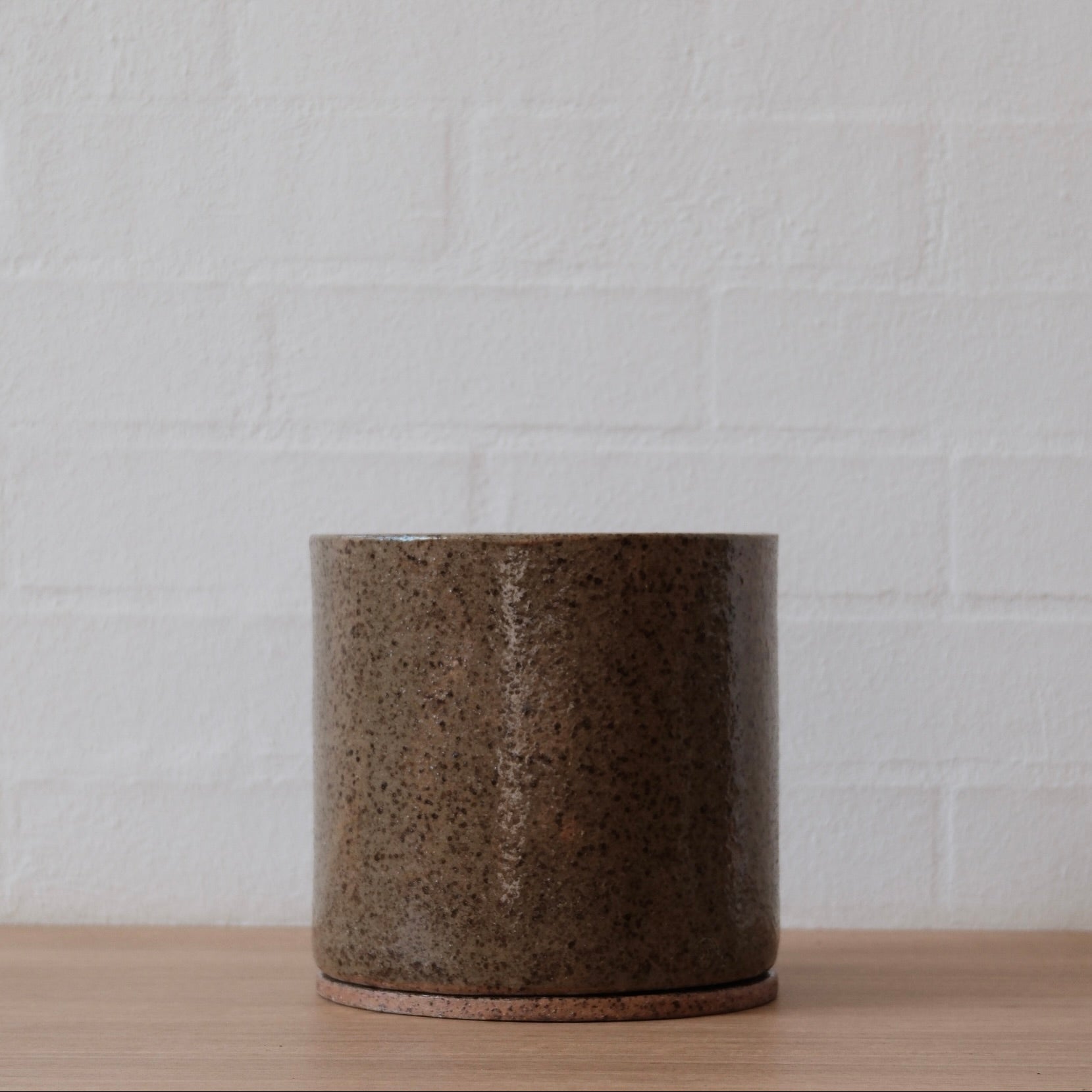 Large cylindrical olive green with speckled ceramic pot on a wooden surface with a white brick wall background.