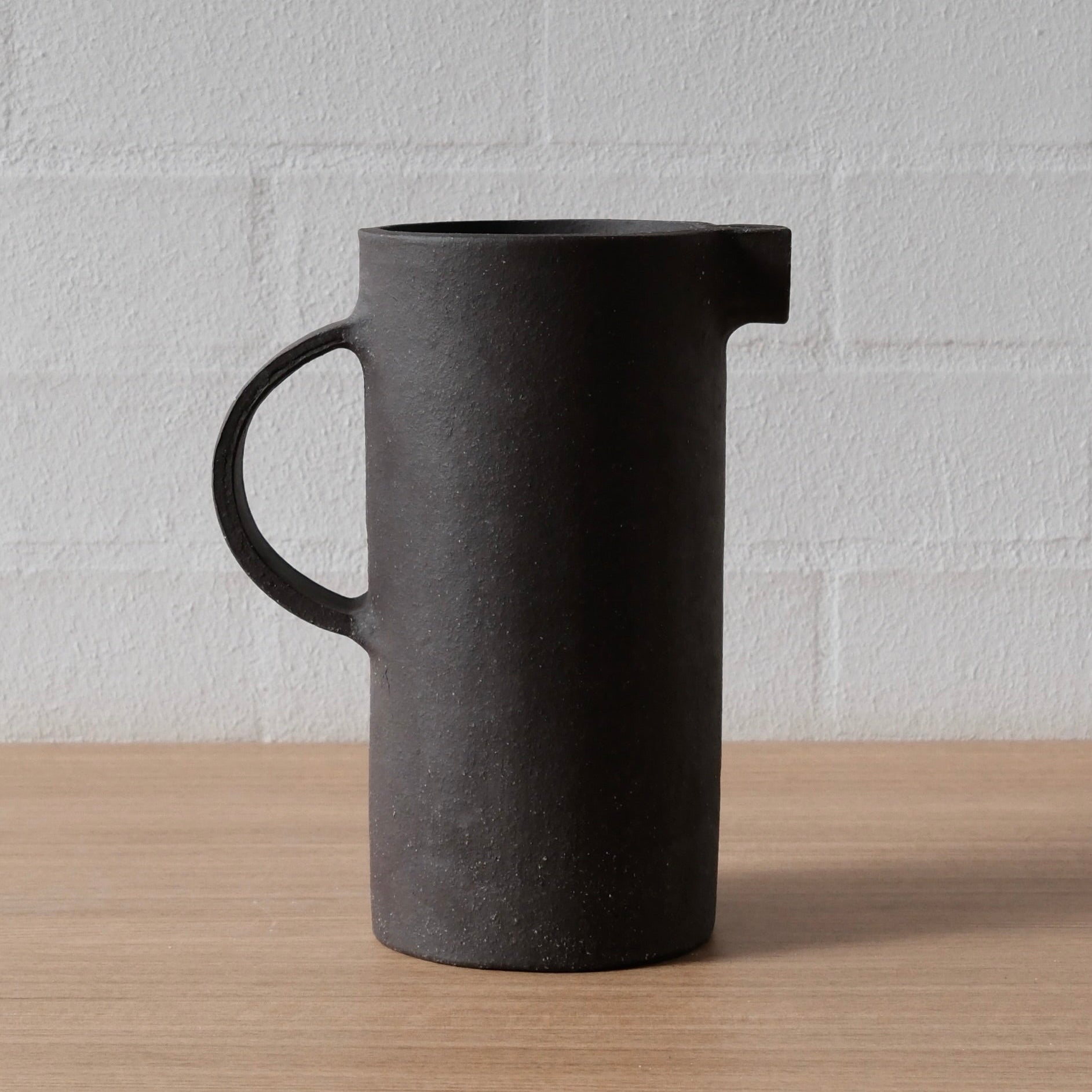 A matte black ceramic pitcher with a C-shaped handle and a small pouring spout, standing on a light wood surface against a white brick wall background.