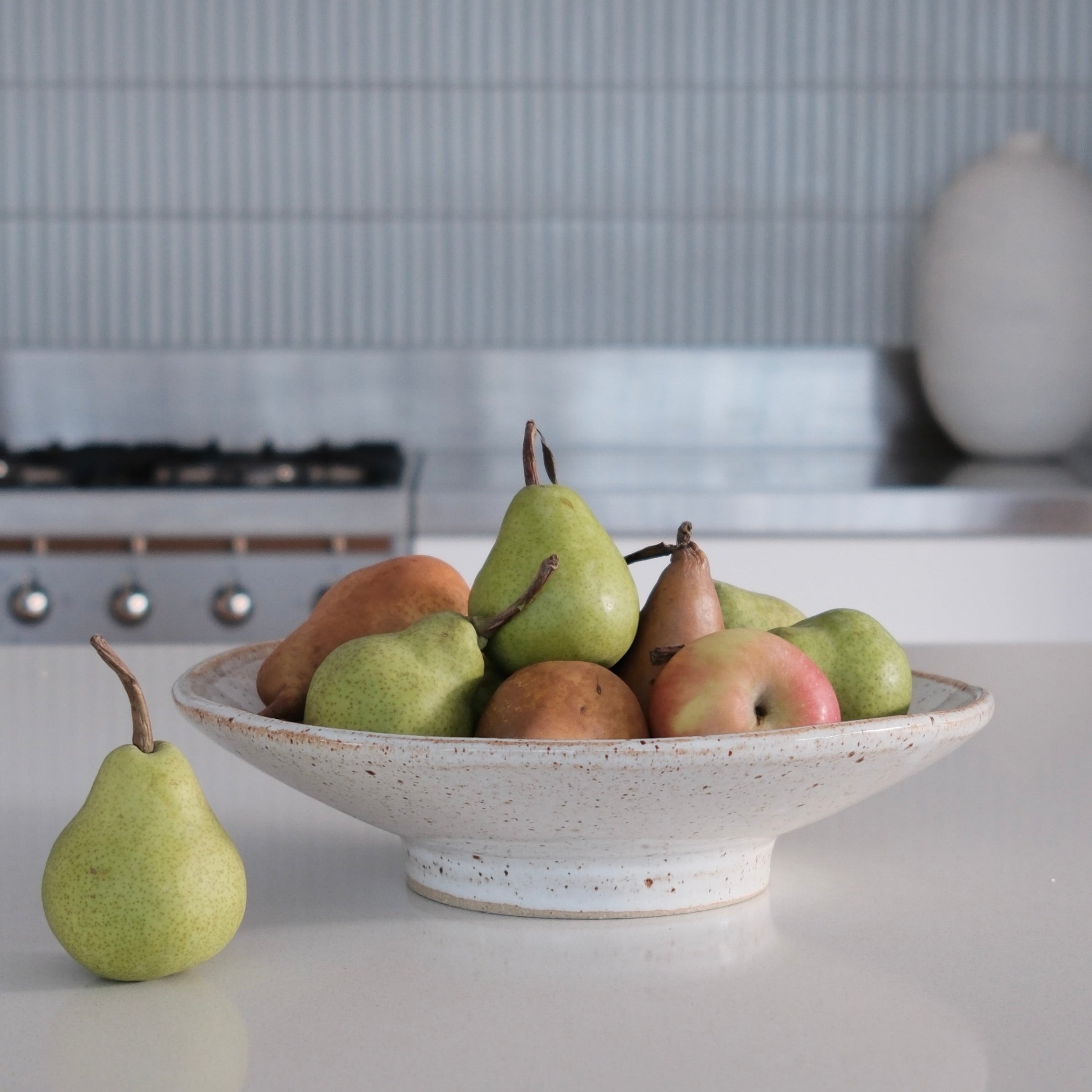 Assorted pears in a ceramic bowl on a kitchen counter with a tiled backsplash.