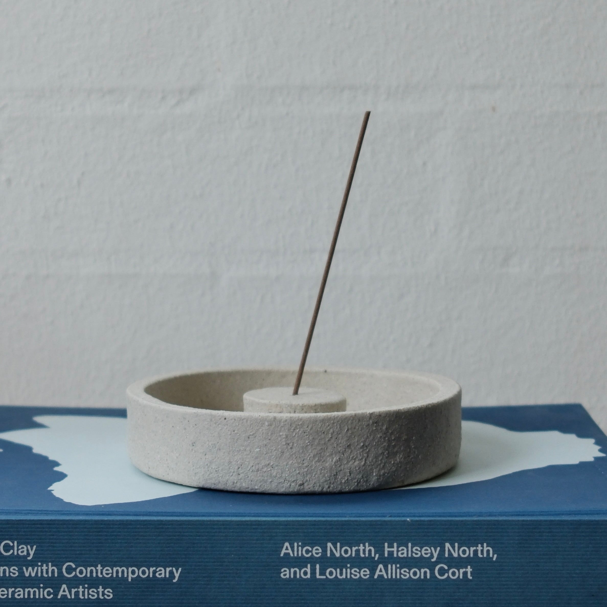 White textured ceramic incense holder with a single incense stick on a book titled 'Earth & Fire'.
