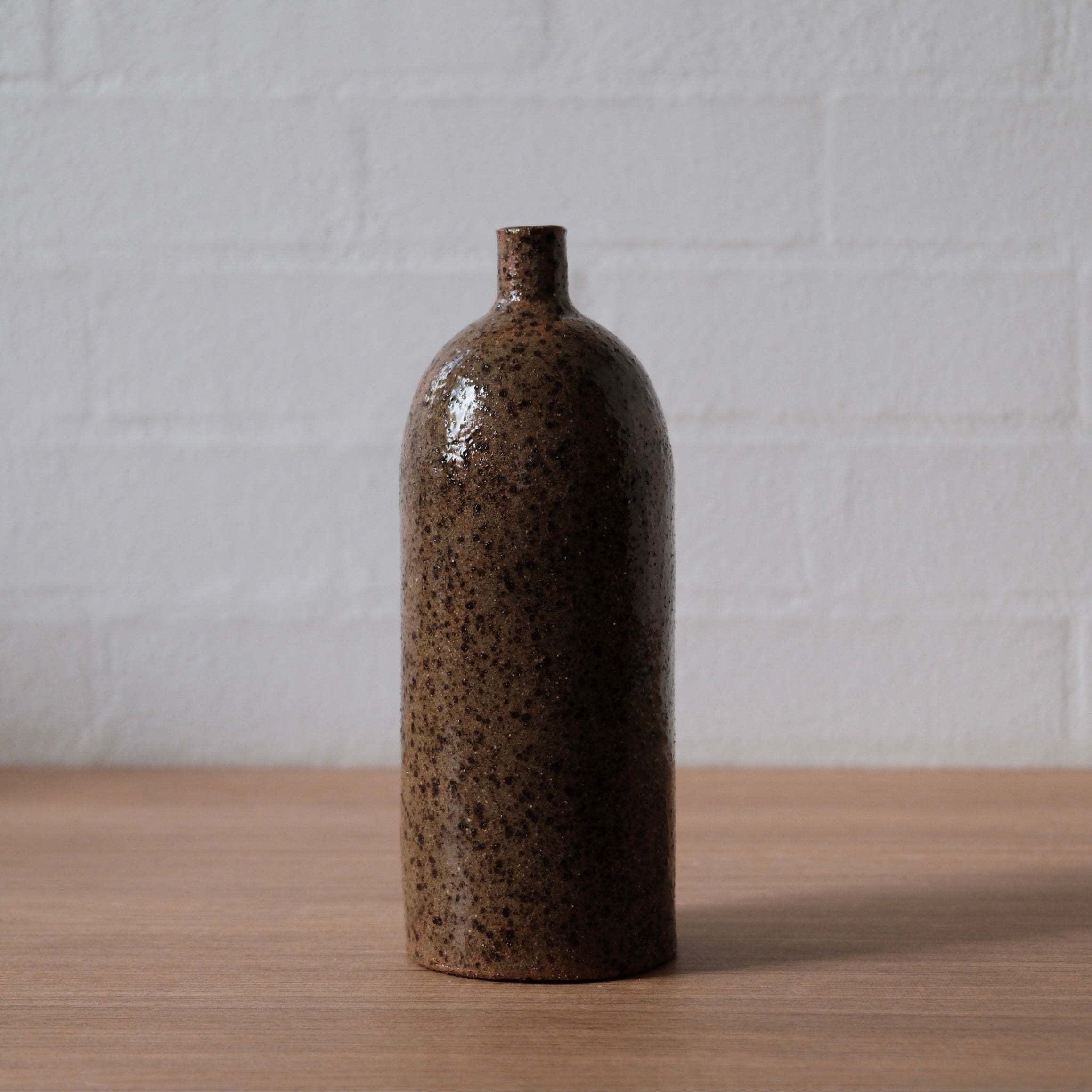 Tall olive speckled ceramic vase, with cylindrical body, rounded top and small spout, one a wooden bench in front of a white wall