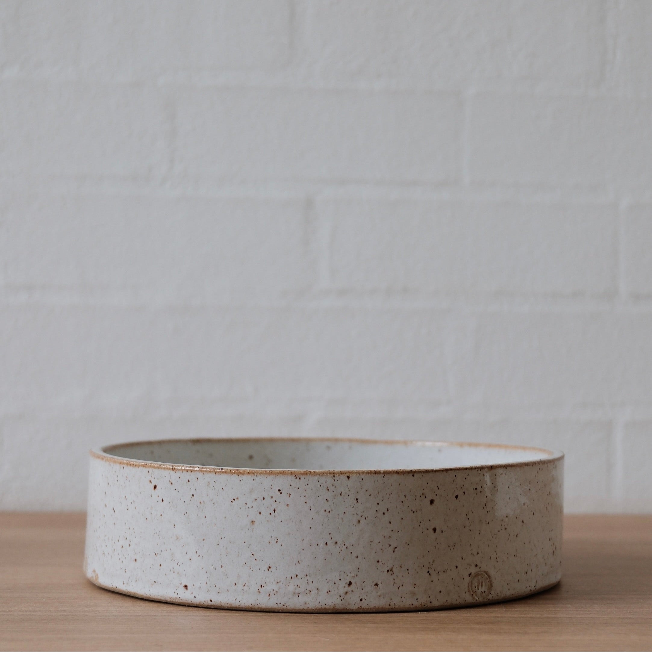 A medium white ceramic bowls with medium straight sides with slightly speckled golden finish on a wooden surface against a white textured wall.