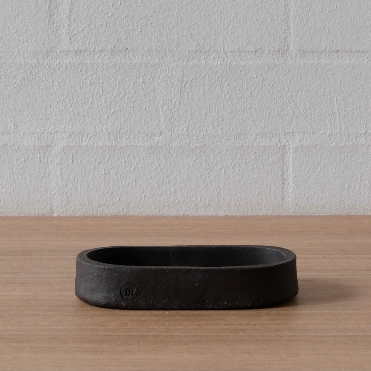 A dark charcoal black ceramic oval-shaped tray or shallow dish with a matte finish, sitting on a light wood surface against a white brick wall background.