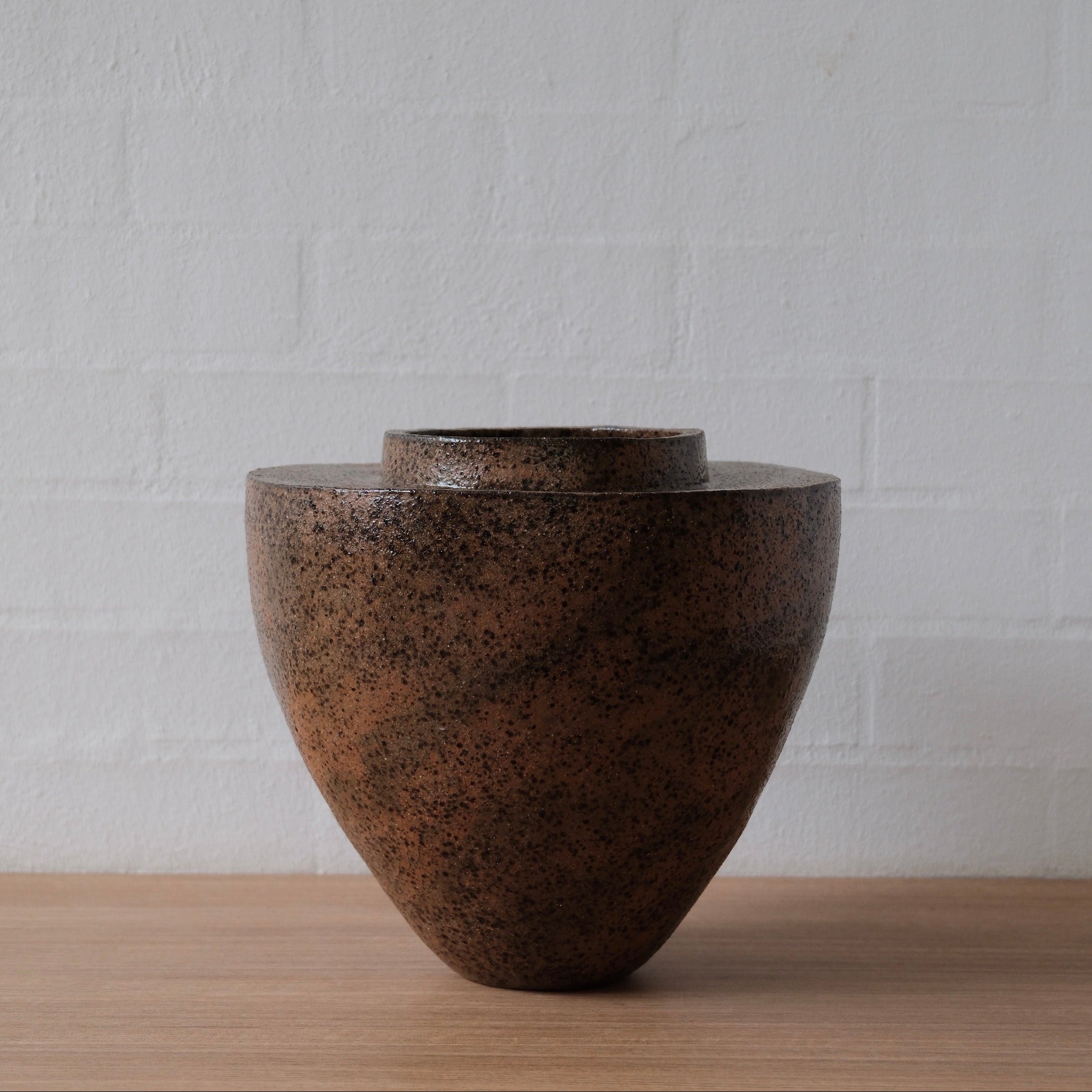 Brown ceramic vessel with narrow base and wide flat top and narrow neck on a wooden surface with a white textured wall background