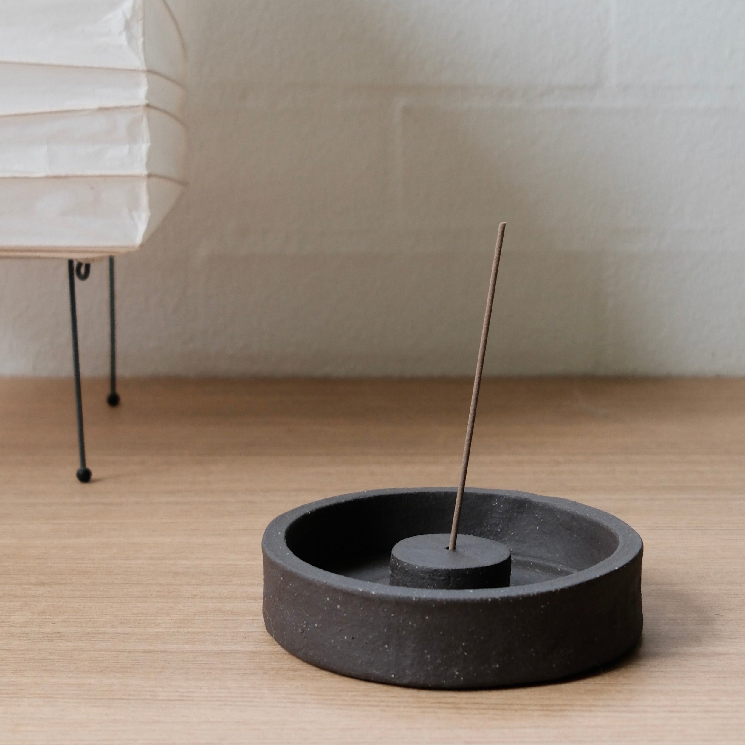 Black ceramic incense holder with a single incense stick on a wooden surface, with a white lamp in the background.