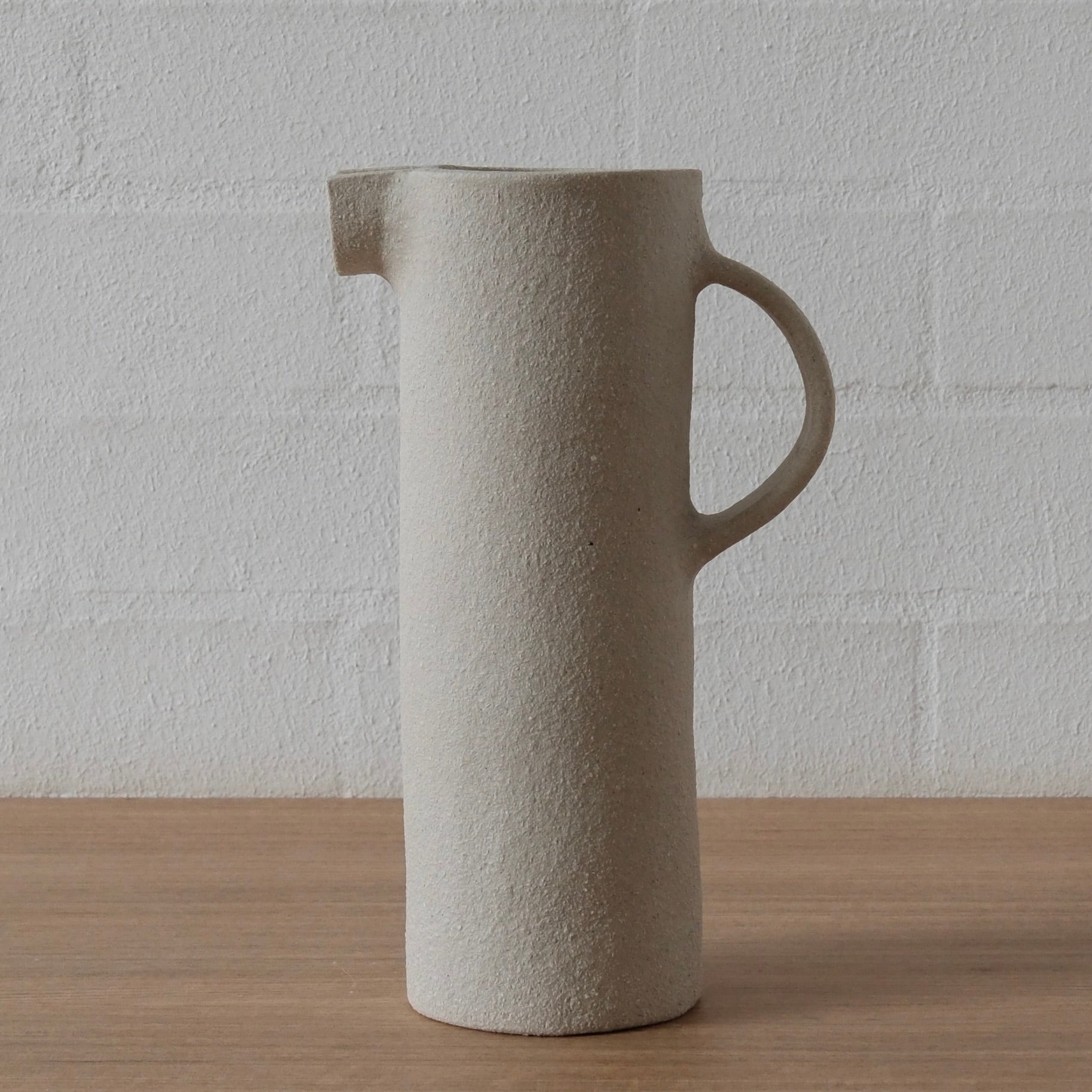 A tall, cylindrical ceramic pitcher with a textured white matte finish, featuring a small pouring spout and a loop handle, set on a light wood surface against a white brick wall.