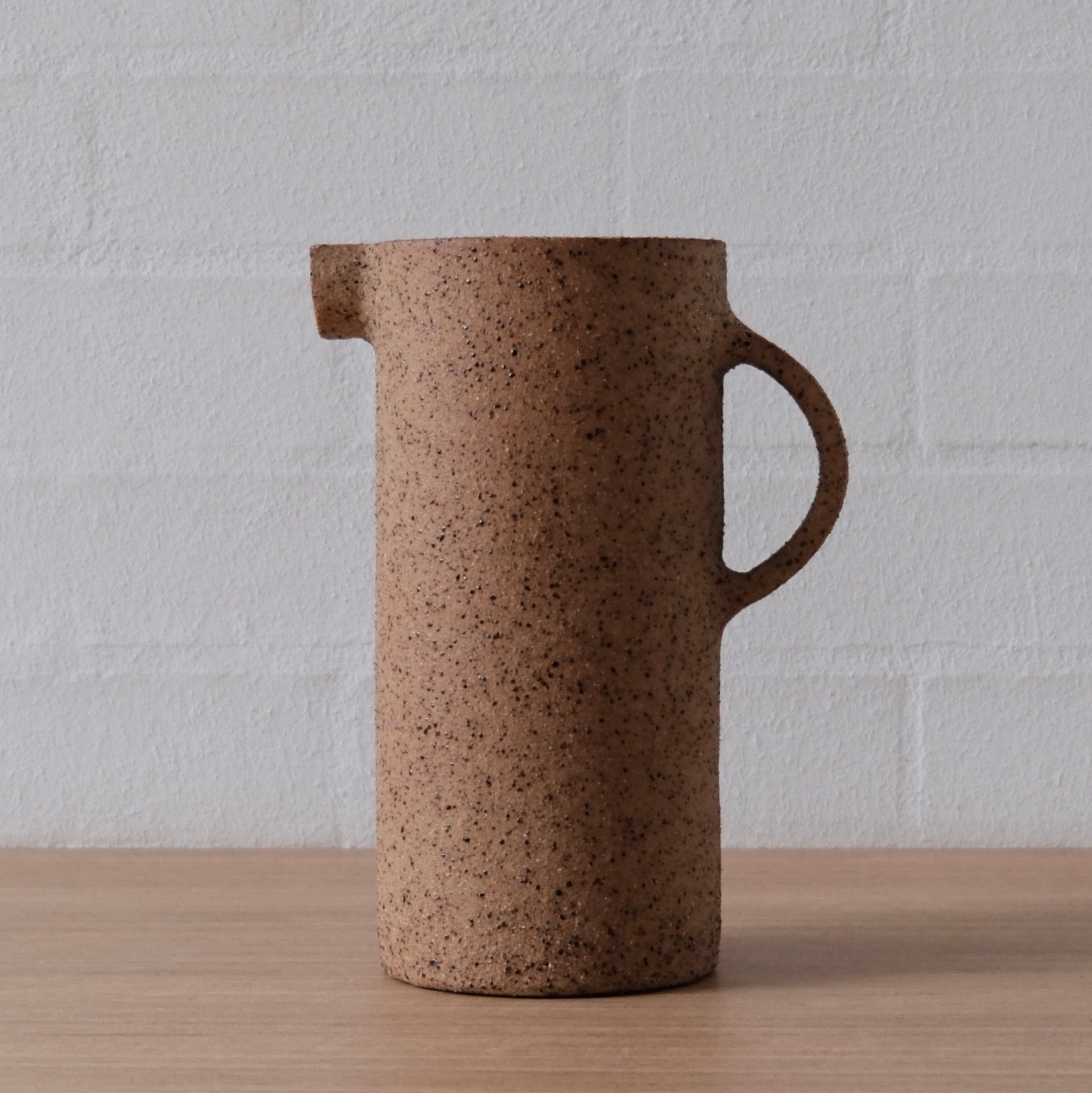 A speckled brown ceramic pitcher with a side handle and pouring spout, standing on a light wood surface against a white brick wall.