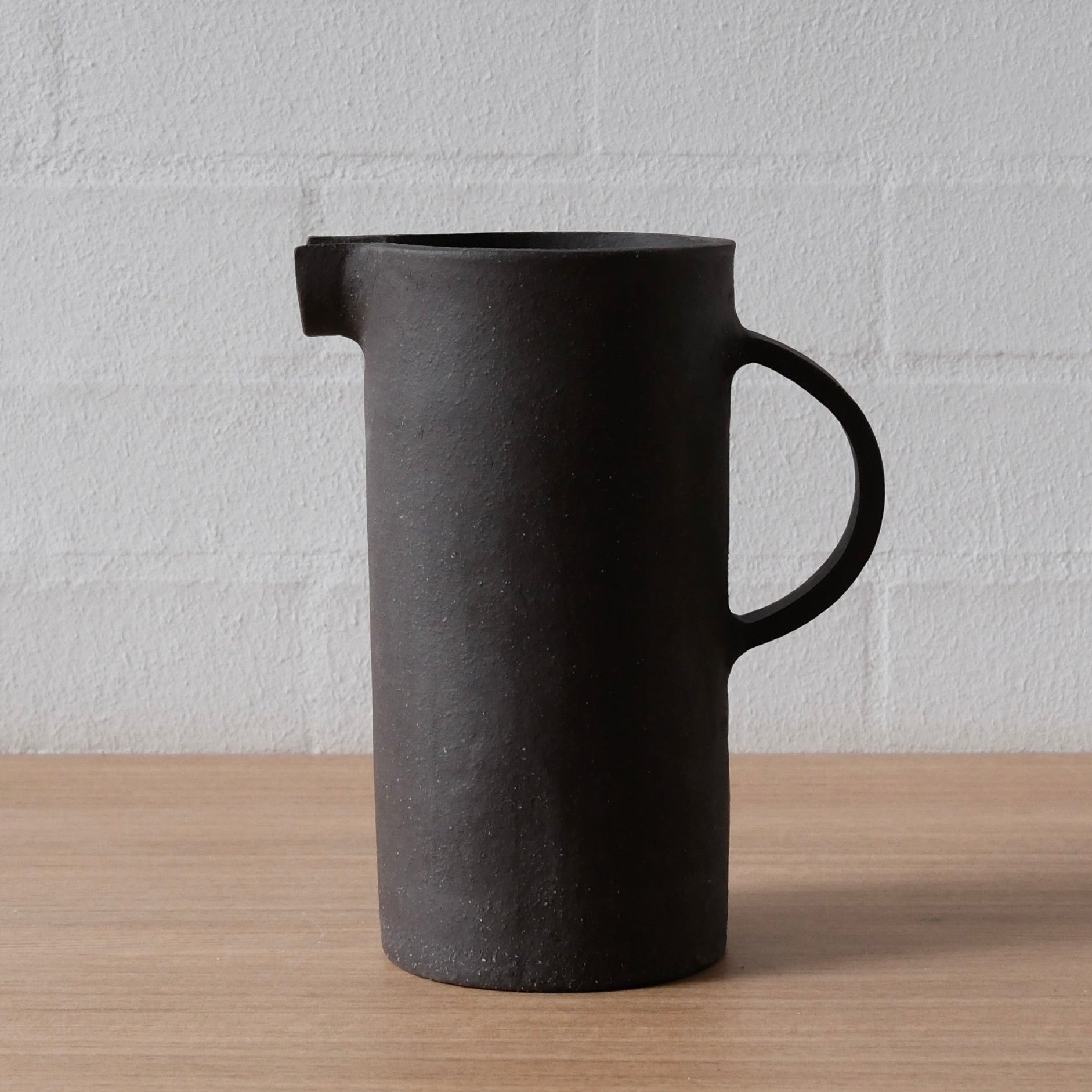A matte black ceramic pitcher with a small integrated spout and a curved handle, standing on a light wood surface against a white brick wall background.