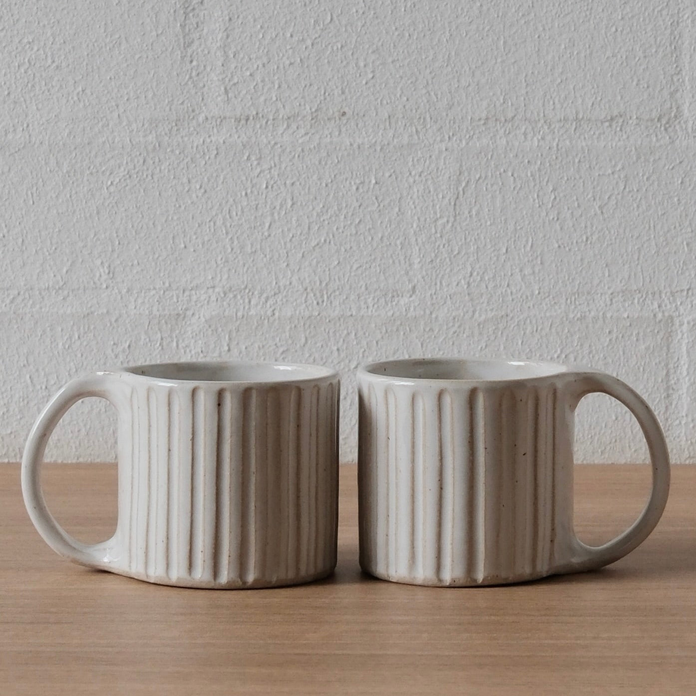 Two ribbed ceramic mugs in a warm off-white glaze, sitting side-by-side on a light wood surface against a white textured brick wall.