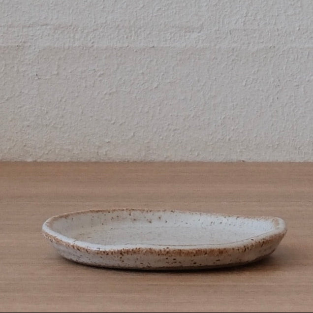 Side view of a small white ceramic plate with slightly speckled finish and golden highlights on a wooden surface and textured white background