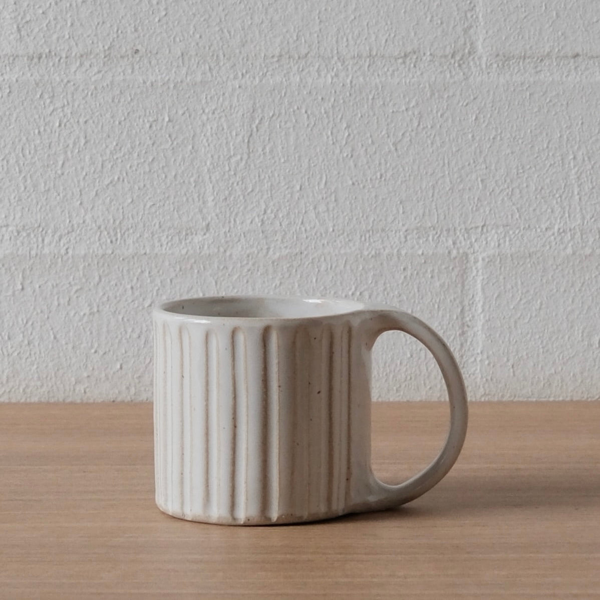 A ceramic mug with a ribbed exterior and a loop handle, sitting on a light wood surface against a white textured brick wall.