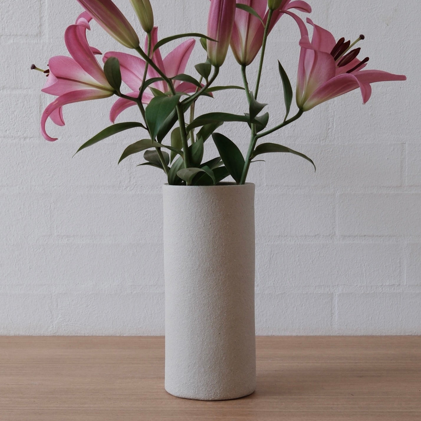Pink lilies arranged in a tall cylindrical white ceramic vase, set on a light wood surface against a white painted brick wall.