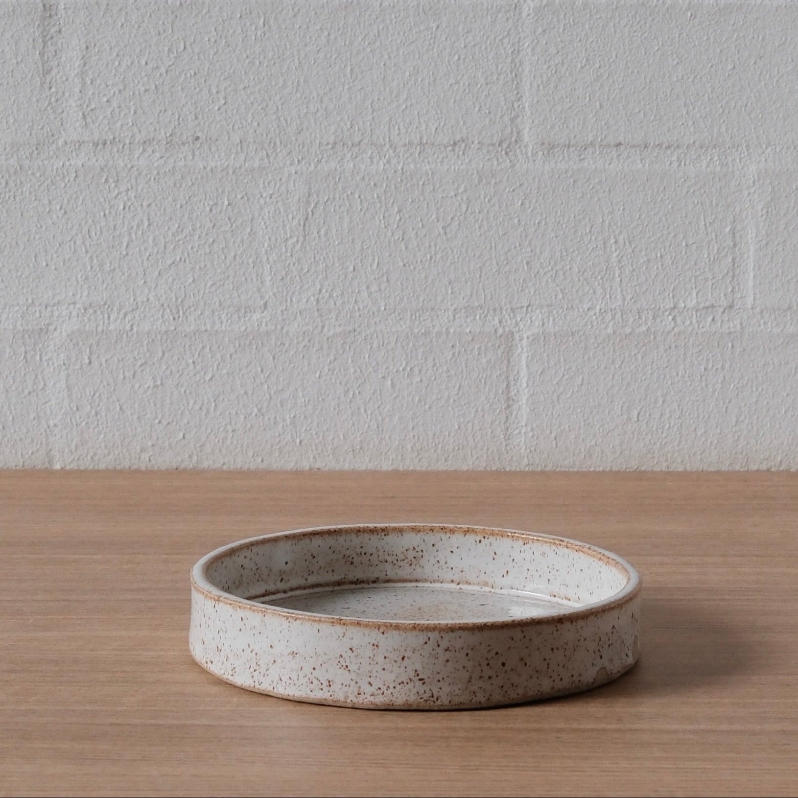 A small round, shallow ceramic tray with a speckled beige and white glaze, sitting on a light wood surface against a white brick wall background.