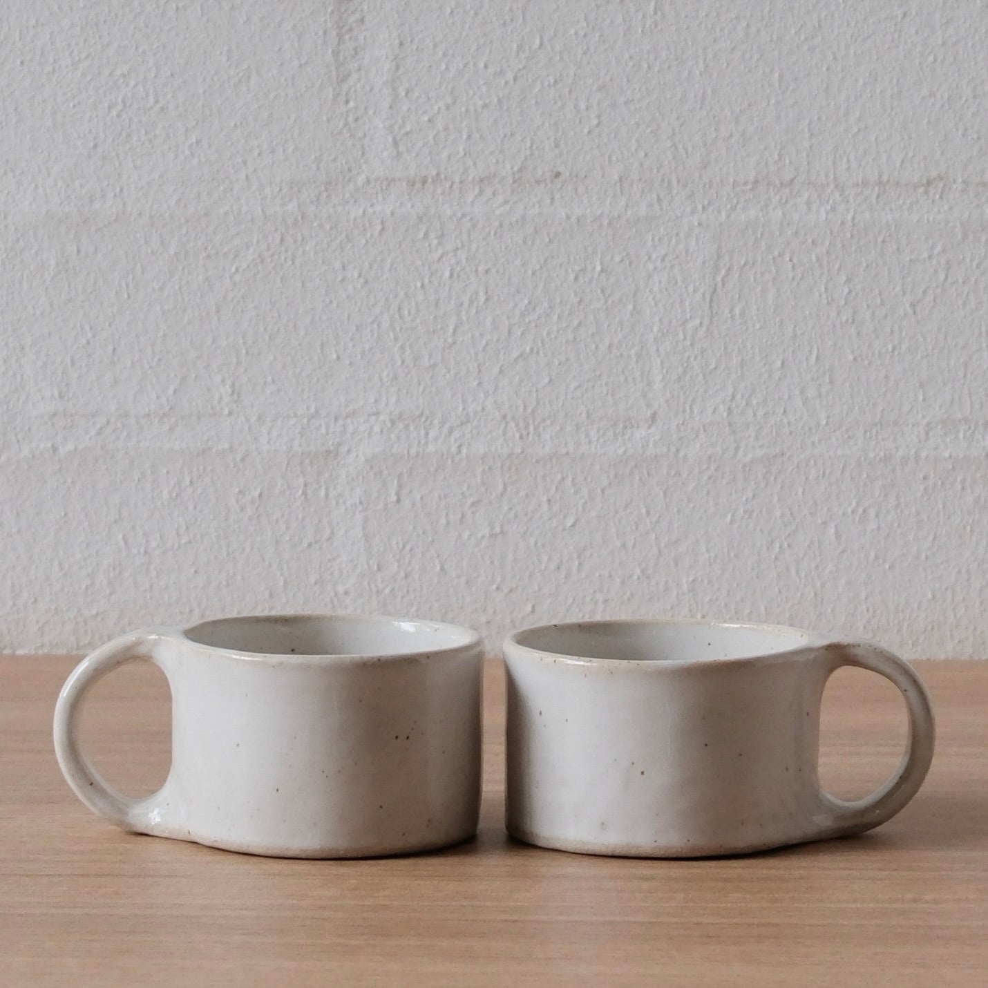 Two small white, slightly speckled ceramic mugs with unique loop handles, sitting side-by-side on a light wood surface against a white brick wall.