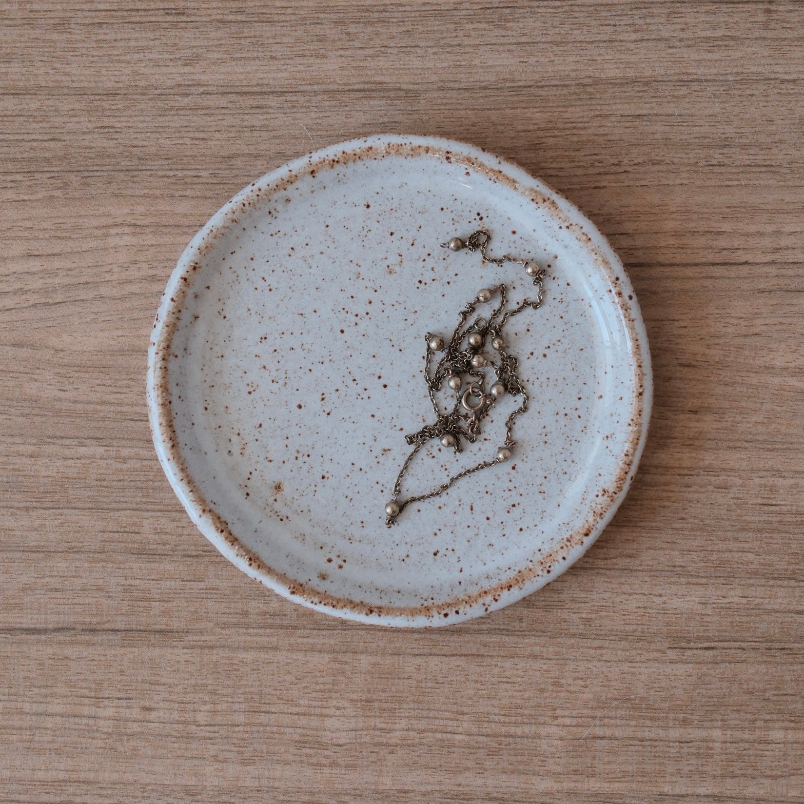 Small white ceramic plate with slightly speckled finish and golden highlights with a gold necklace on a wooden surface