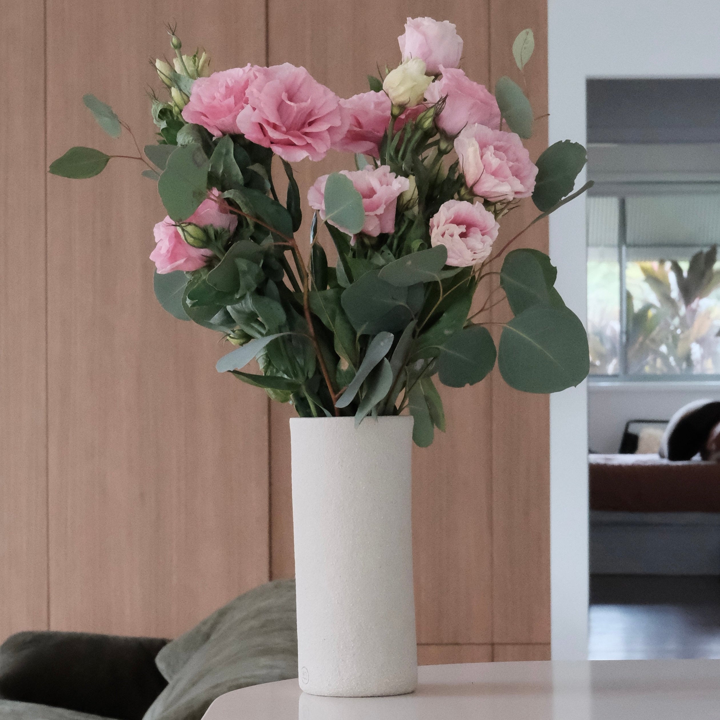 A bouquet of pink roses and green eucalyptus leaves arranged in a tall, cylindrical white ceramic vase, set on a light-colored tabletop in a modern interior setting.