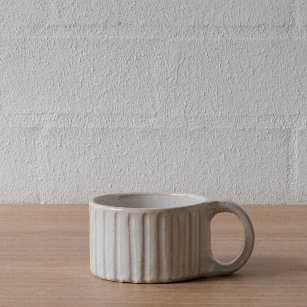 A small ceramic mug with a ribbed exterior and a loop handle, sitting on a light wood surface against a textured white brick wall.