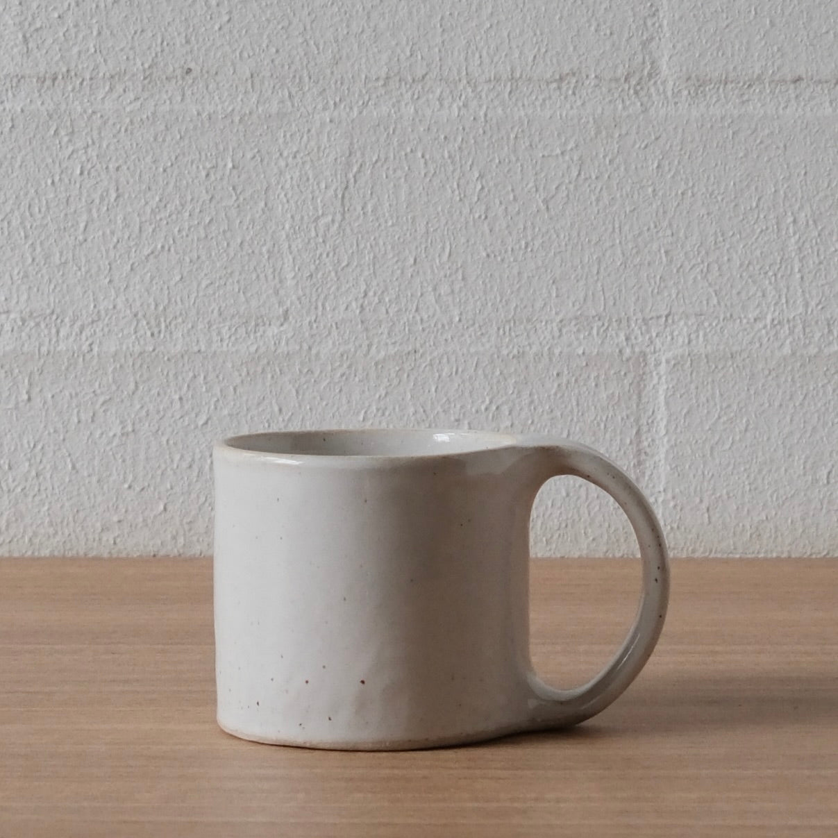 A handmade ceramic mug with a large loop handle, sitting on a light wood tabletop against a white brick wall.