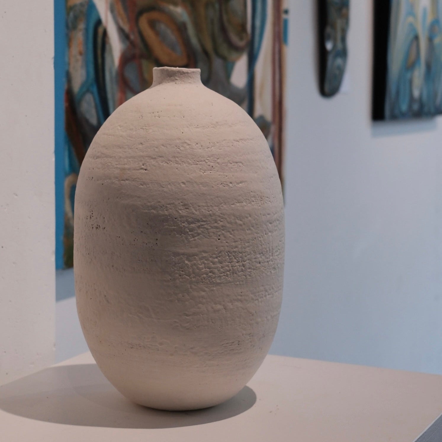 Textured white ceramic vessel on a plinth with abstract art in the background