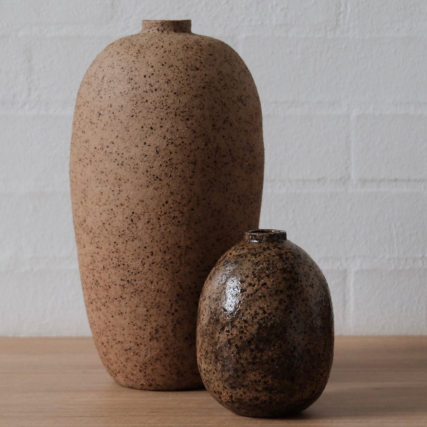 Two ceramic vessels with speckled textures, one shiny and one matte, on a wooden surface and white background