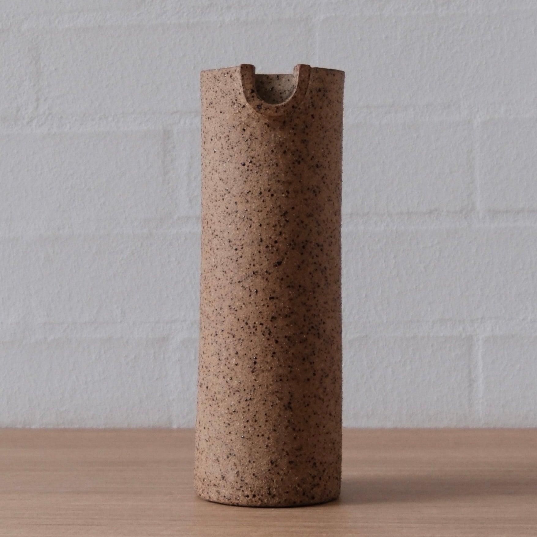 A cylindrical ceramic pitcher with a speckled brown finish, featuring a small pouring spout, standing on a light wood surface against a white brick wall.