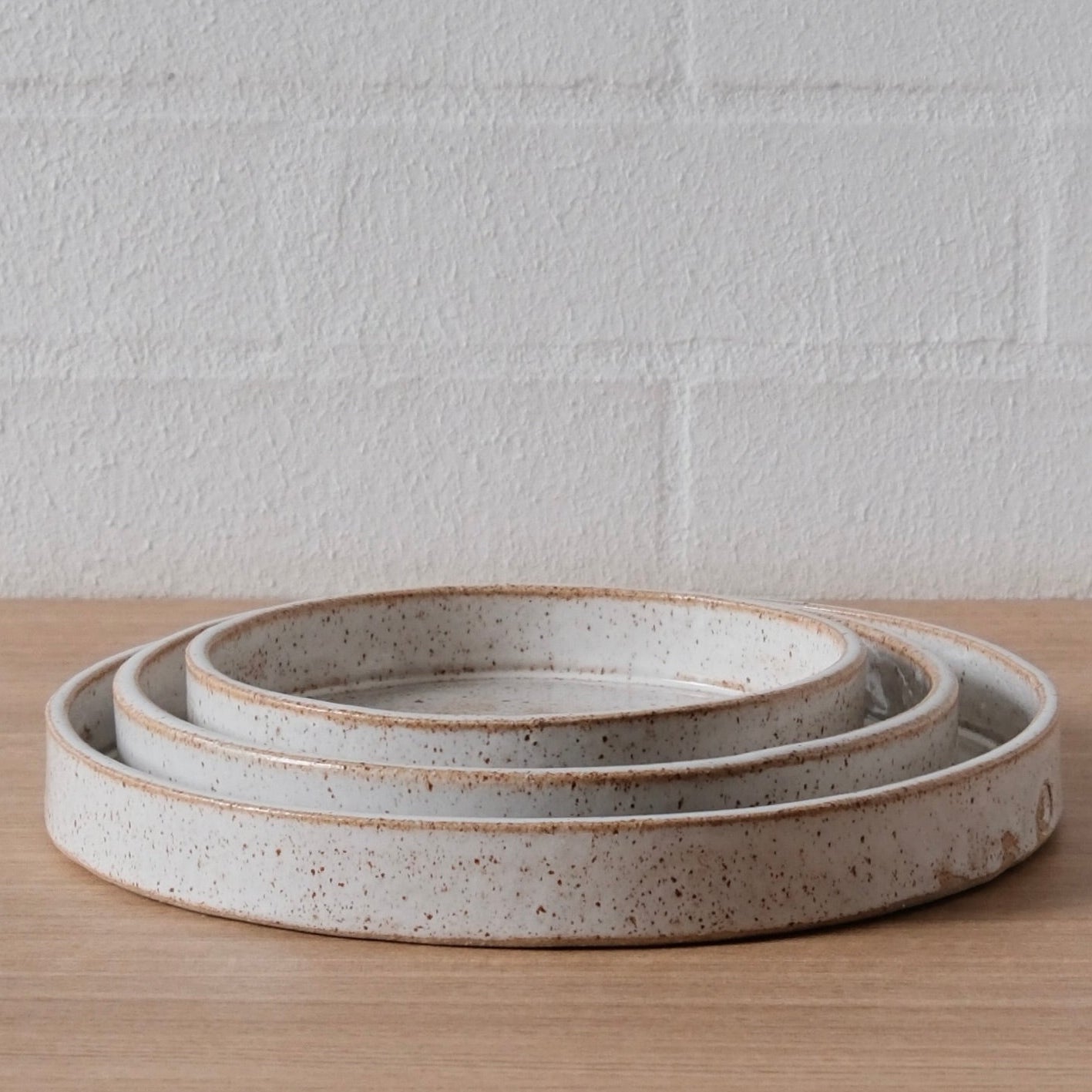 Three nested round ceramic trays with a speckled off-white glaze and a rustic brown rim, displayed on a light wood surface against a white brick wall.