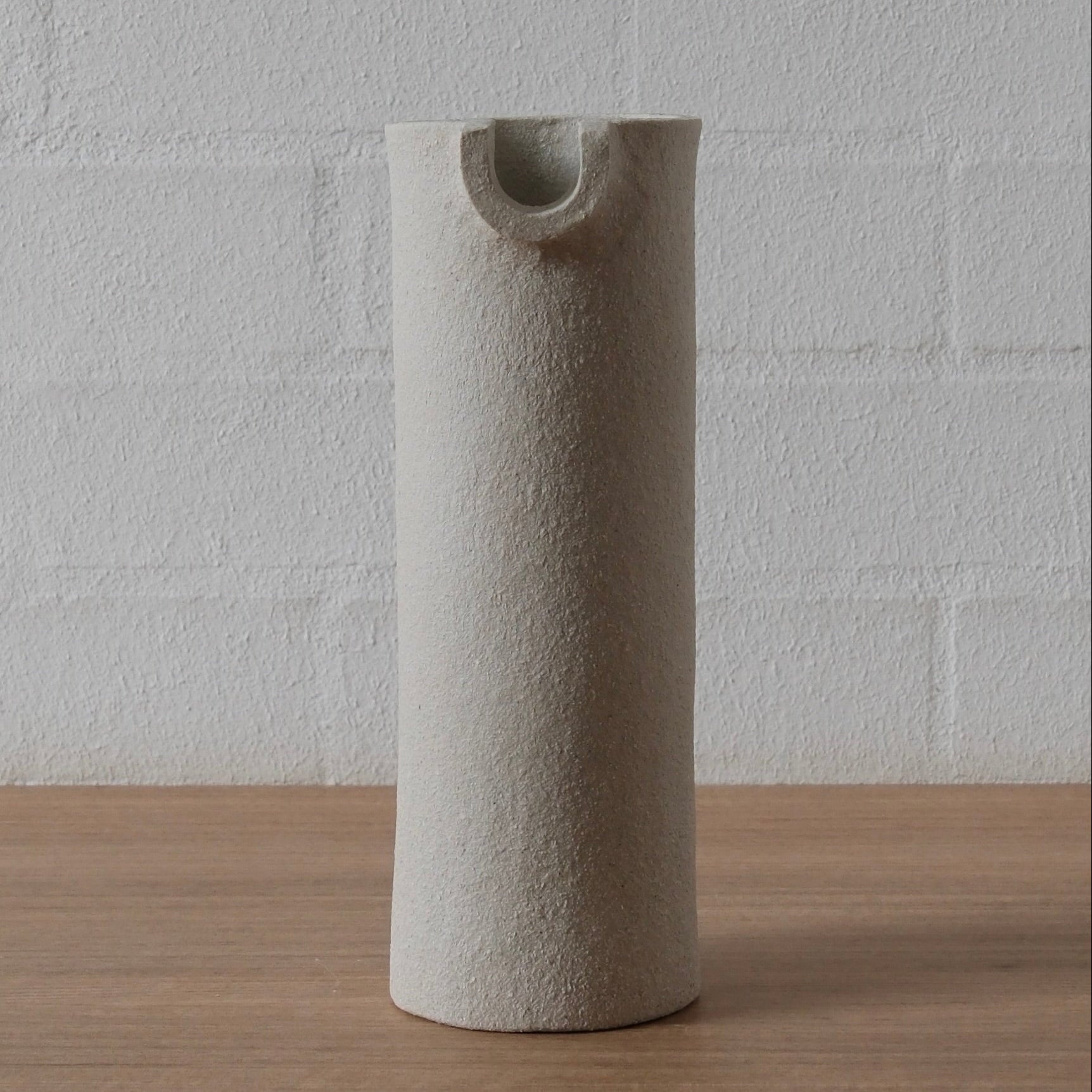 A tall, cylindrical white textured ceramic vessel with a small integrated opening near the top, standing on a light wood surface against a white painted brick wall.