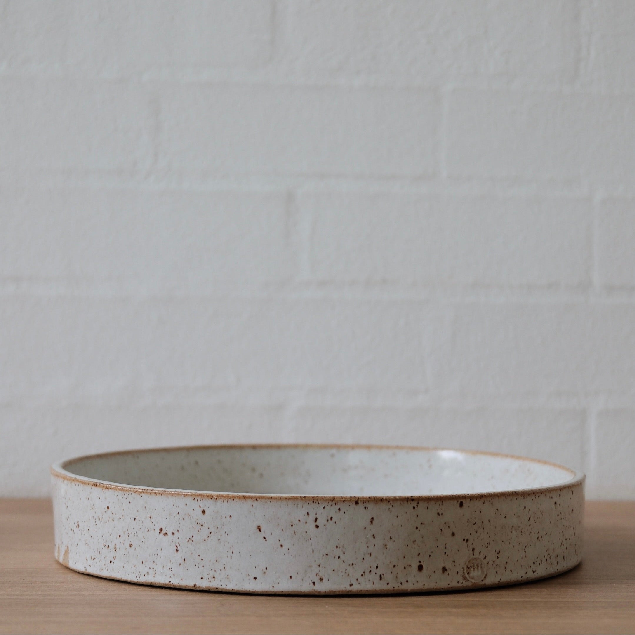 A large white ceramic bowls with low straight sides with slightly speckled golden finish on a wooden surface against a white textured wall.