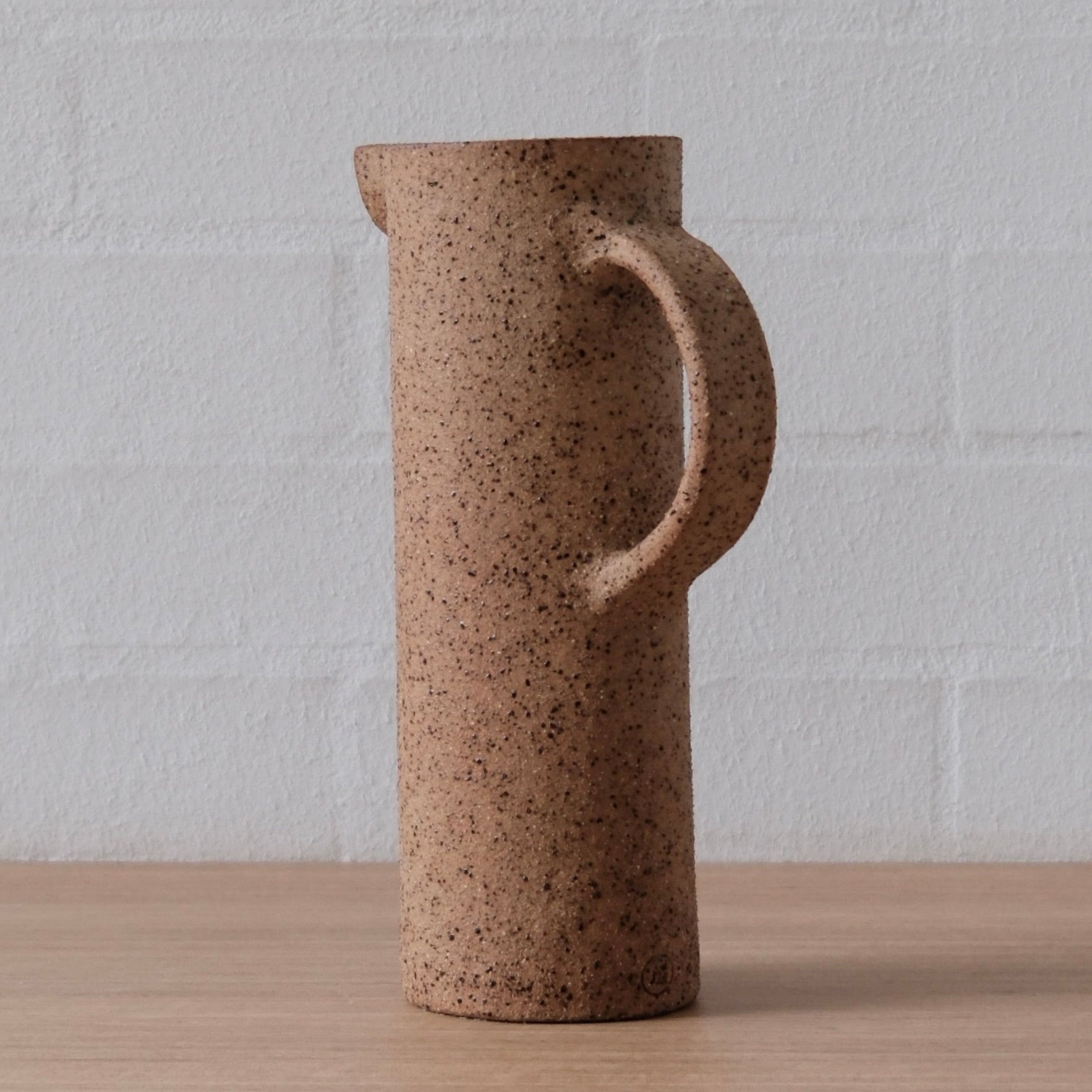 A tall cylindrical ceramic pitcher with a speckled brown finish, featuring a curved handle and a small pouring lip, standing on a light wood surface against a white brick wall.