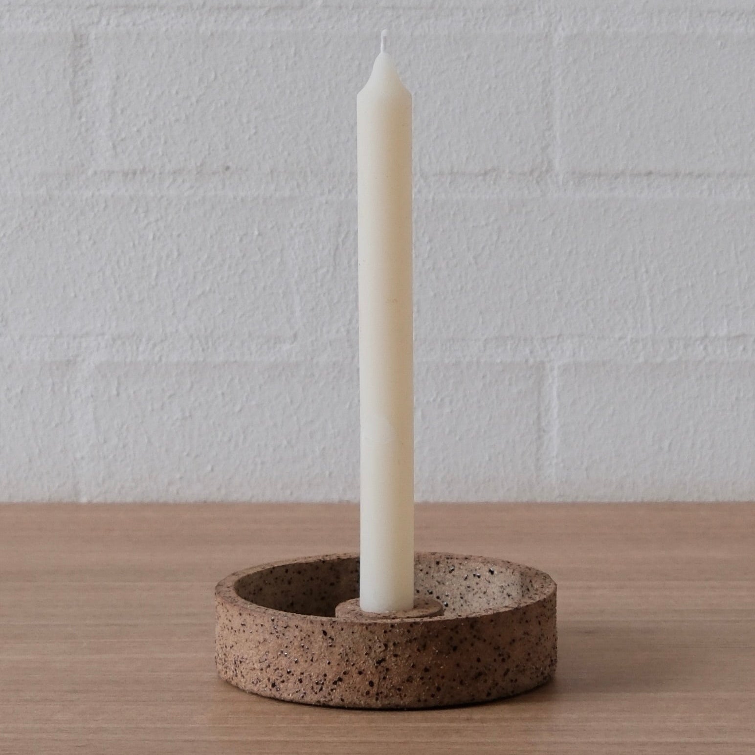 A white candle standing upright in a round, speckled brown ceramic holder on a light wood surface against a white brick wall background.