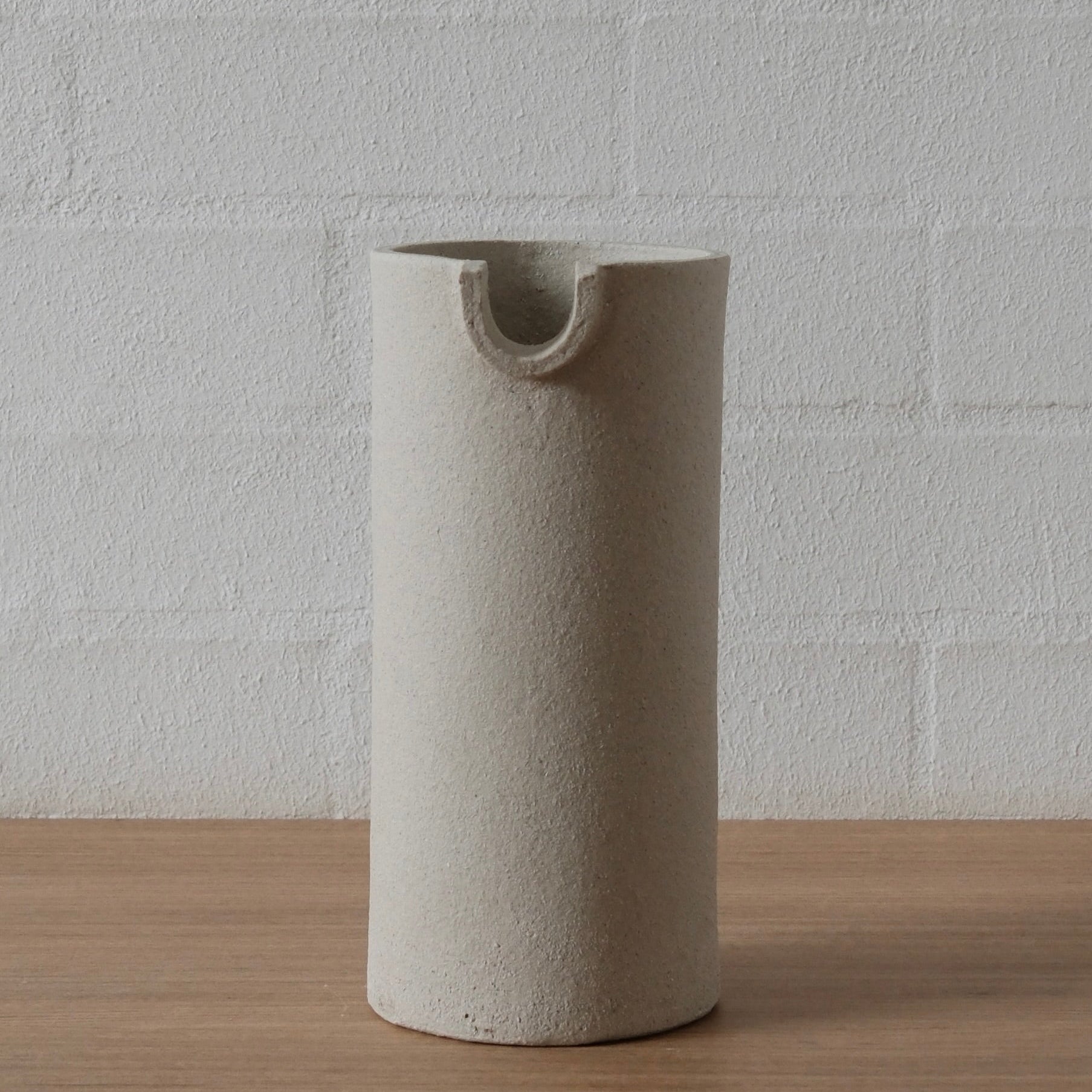 A minimalist, cylindrical ceramic pitcher or vase with a small integrated loop at the rim, standing on a light wood surface against a white painted brick wall.