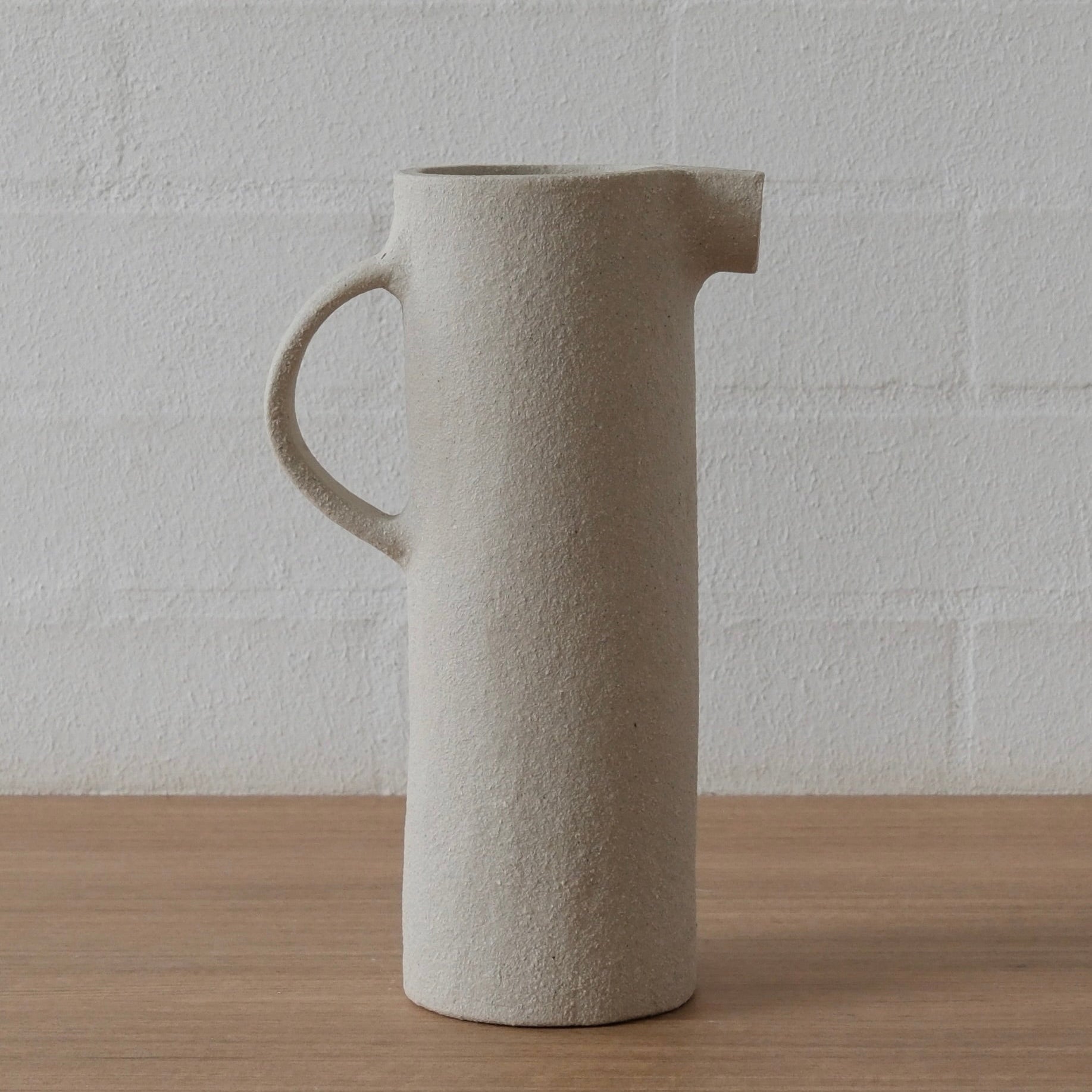 A tall, cylindrical ceramic pitcher with a textured white matte finish, featuring a side handle and a small pouring spout, set on a light wood surface against a white brick wall.