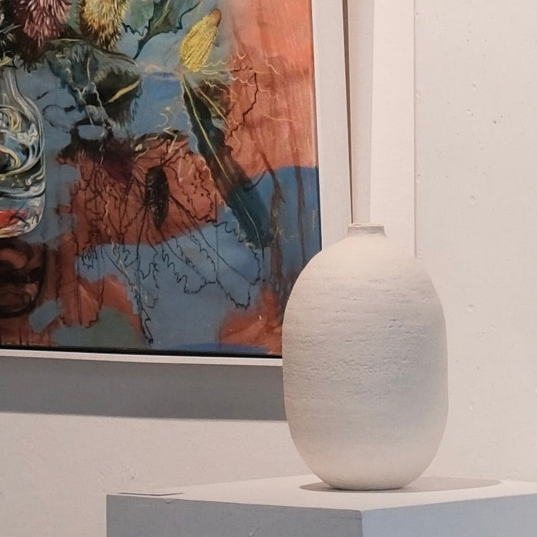 Framed painting of a still life with flowers on a white wall, accompanied by a lRGE white vessel on a plinth.