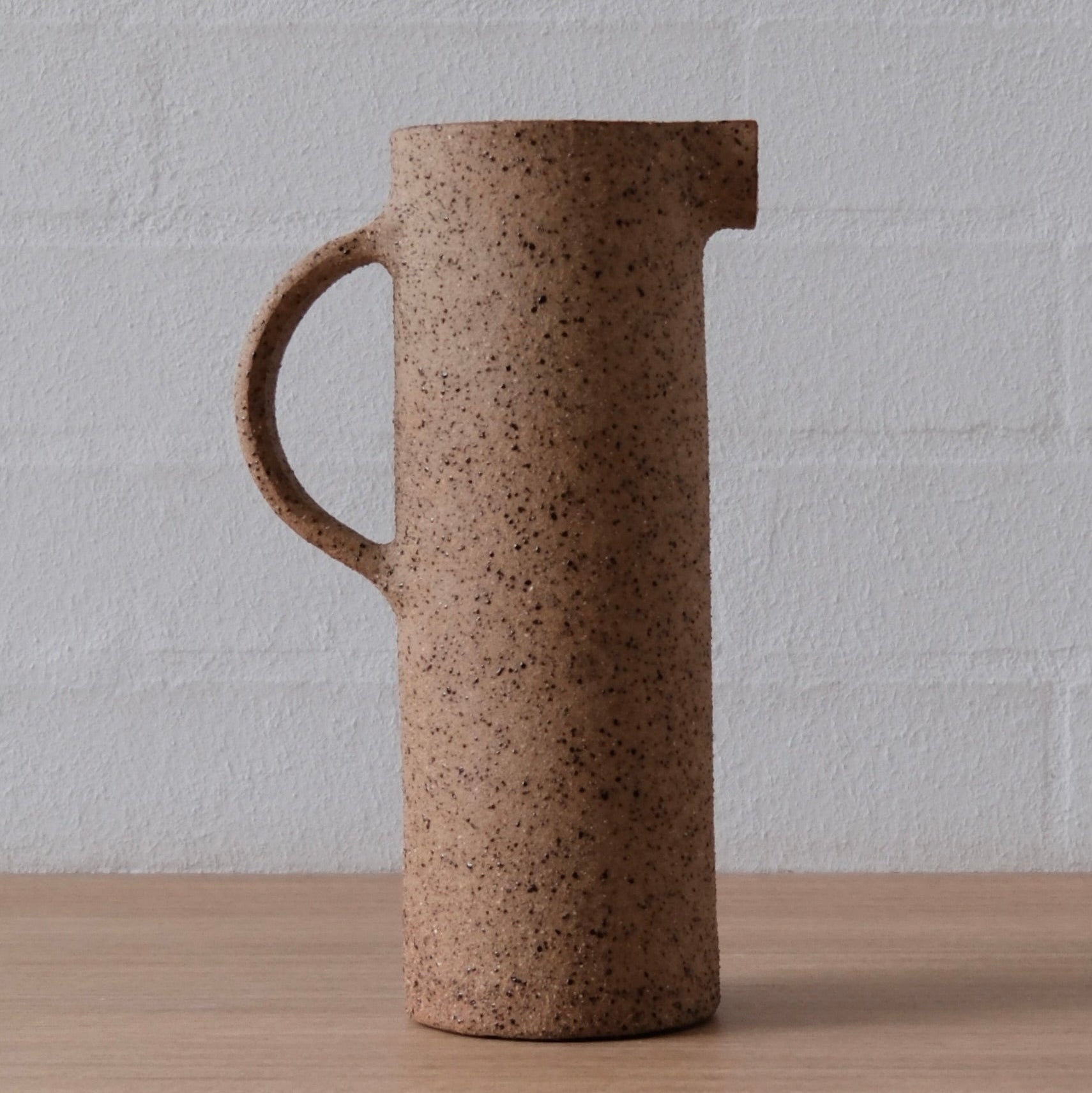 A tall cylindrical ceramic pitcher with a handle and pouring spout, featuring a speckled brown stoneware finish, set on a light wood surface against a white brick wall.