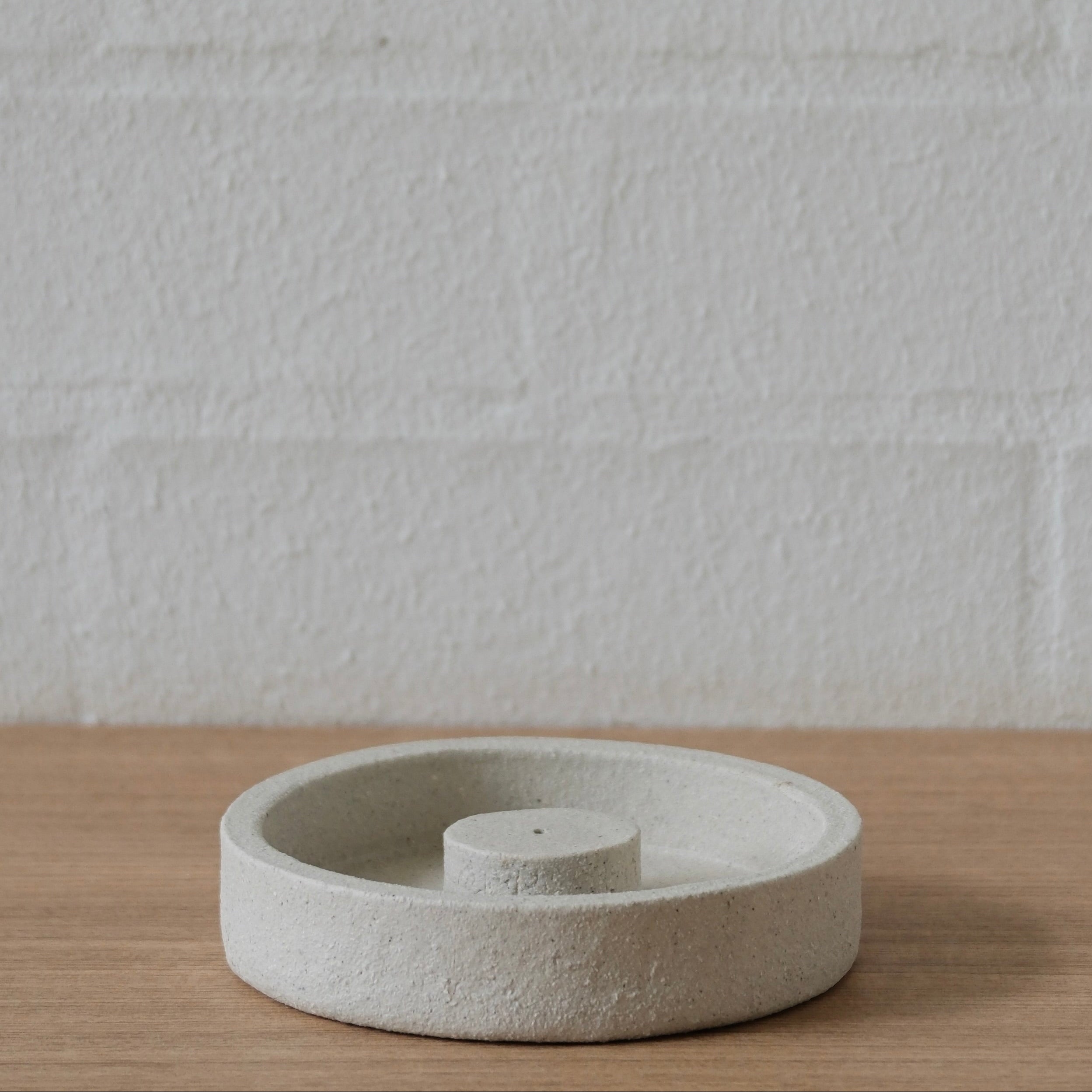Textured, round white ceramic incense holder on a wooden surface with a white wall background