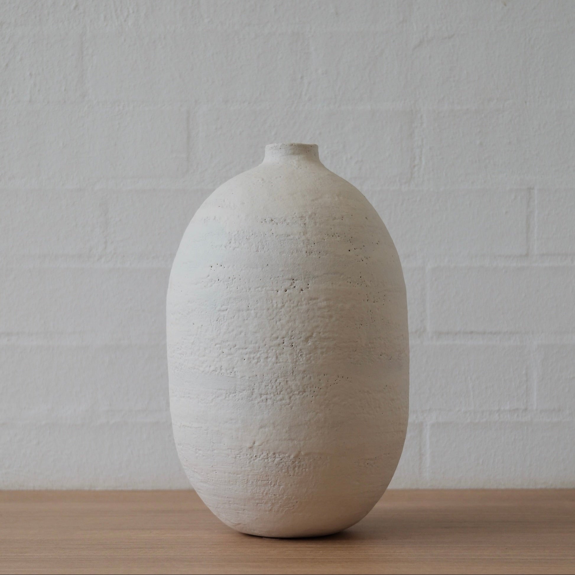 Large, oval white textured vessel on a wooden surface with a white brick wall background
