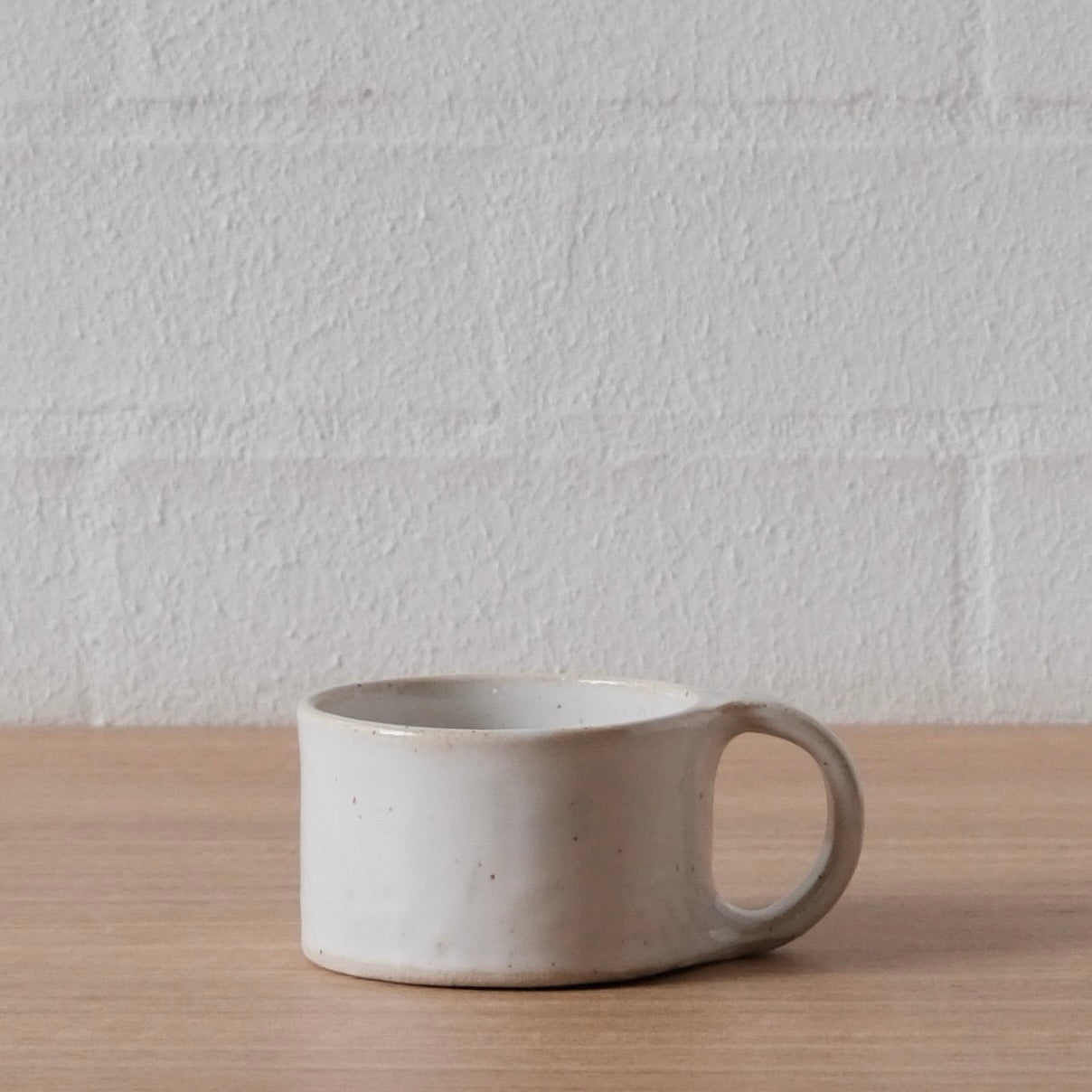 A small handmade ceramic mug with a cylindrical body and a loop handle, sitting on a light wood tabletop against a white brick wall background.