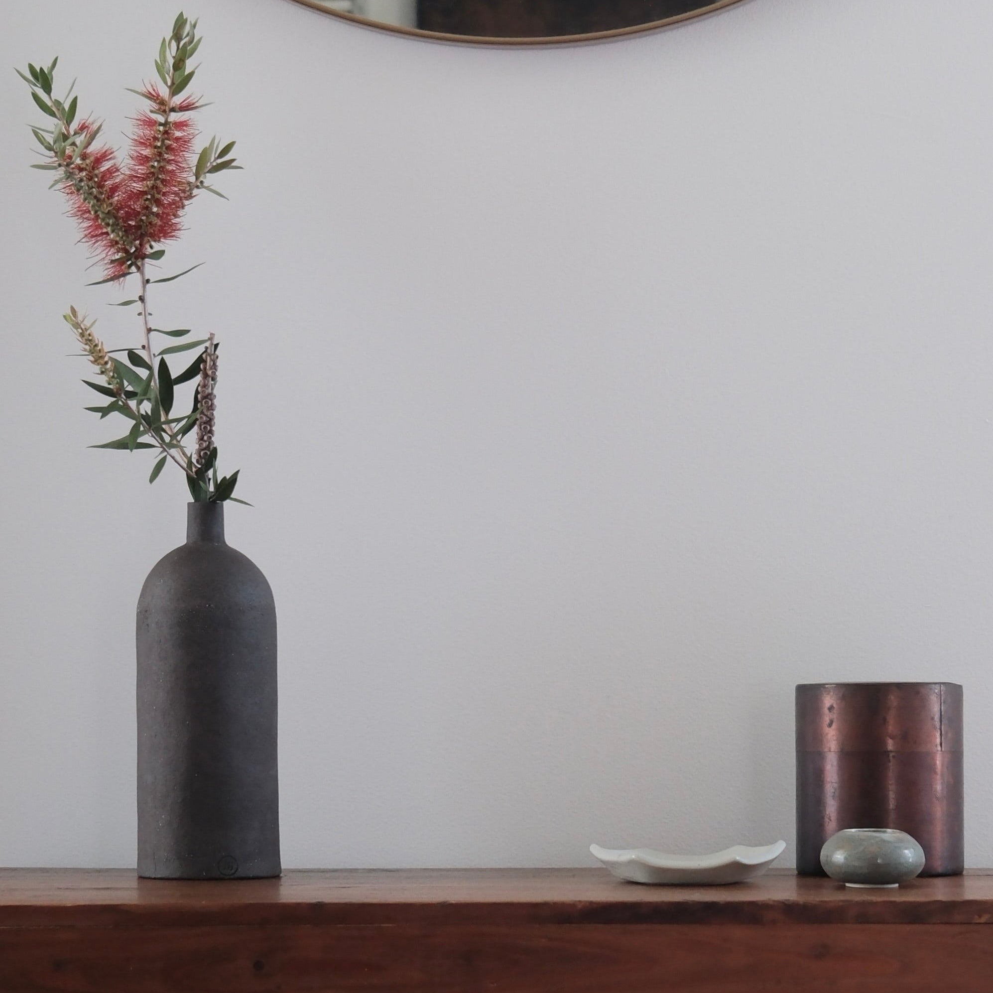 A matte black ceramic bottle vase holding pink and green dried flowers, styled on a dark wood console table against a white wall with a copper candle holder and a small dish.