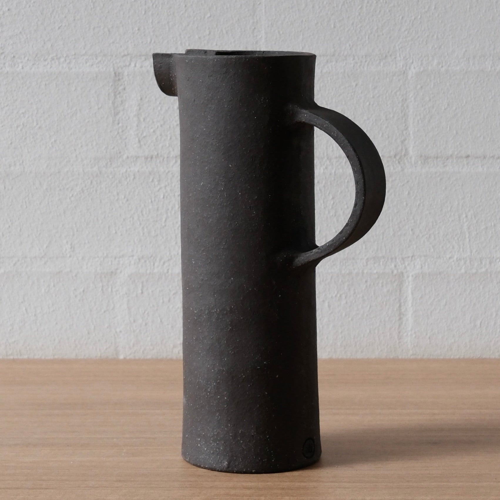 A matte black ceramic pitcher with a cylindrical body, integrated handle, and small pouring spout, standing on a light wood surface against a white brick wall.