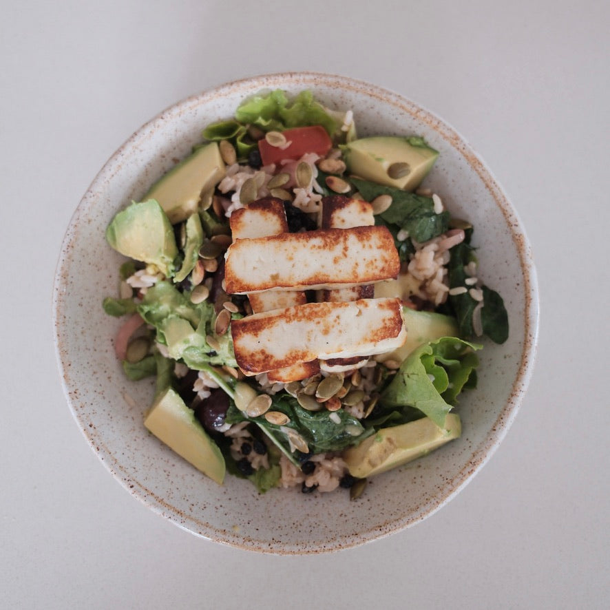 A top-down view of a prepared salad in a speckled white bowl, featuring grilled halloumi, avocado slices, mixed greens, and seeds on a light neutral background.