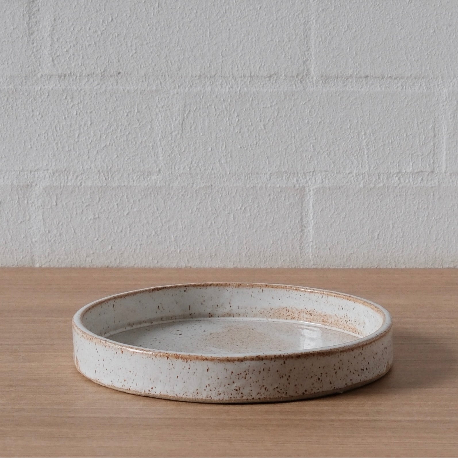 A medium round ceramic tray with a speckled off-white glaze and a rustic brown rim, resting on a light wood surface against a white brick wall.