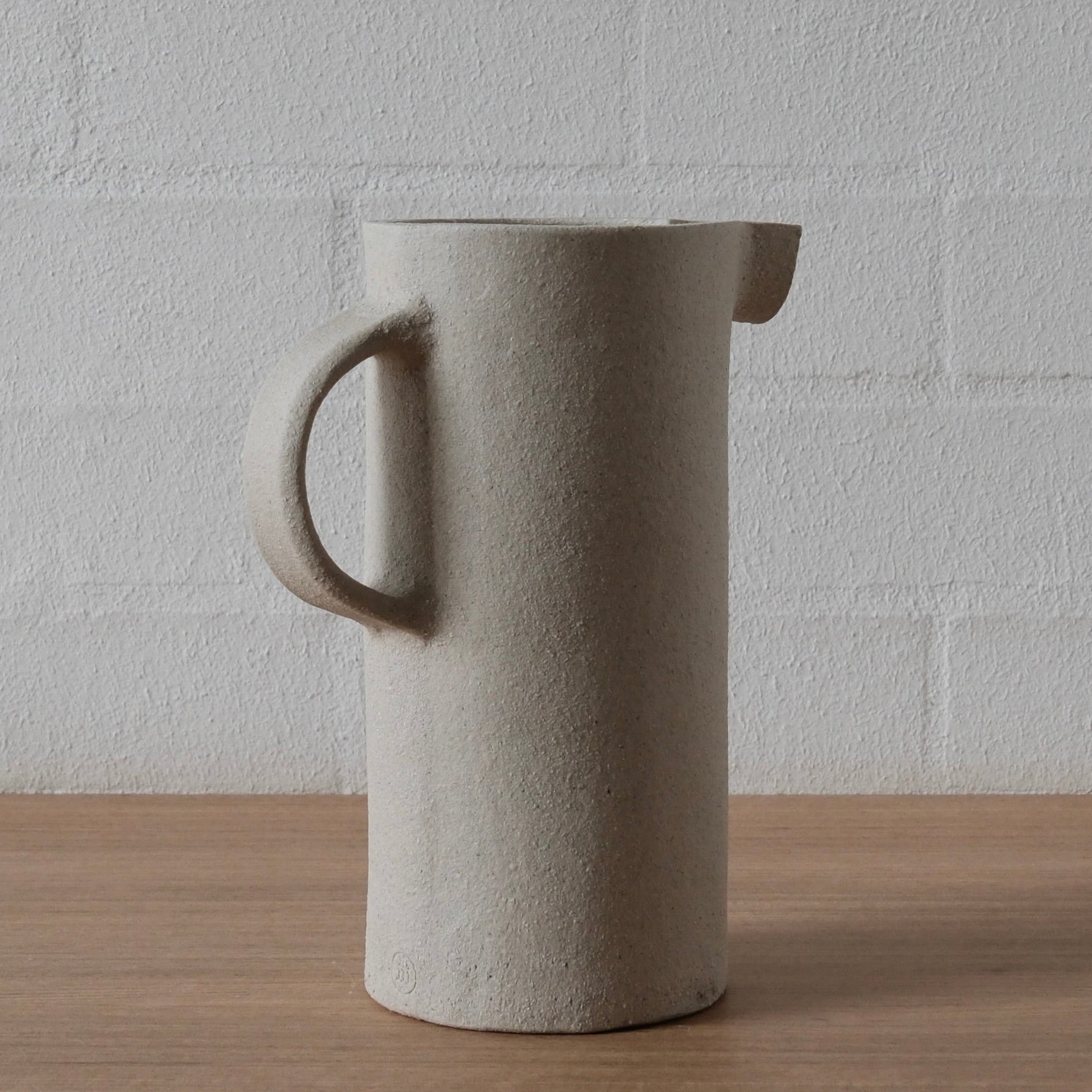 A tall cylindrical ceramic pitcher with a textured white matte finish, featuring a loop handle on the left and a small pouring spout on the right, set on a wooden surface against a white brick wall.