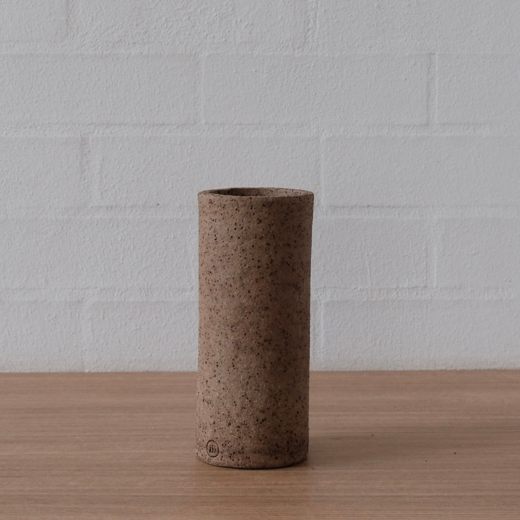 A cylindrical ceramic vase with a textured, speckled earthy brown finish, standing on a light wood surface against a white brick wall background.