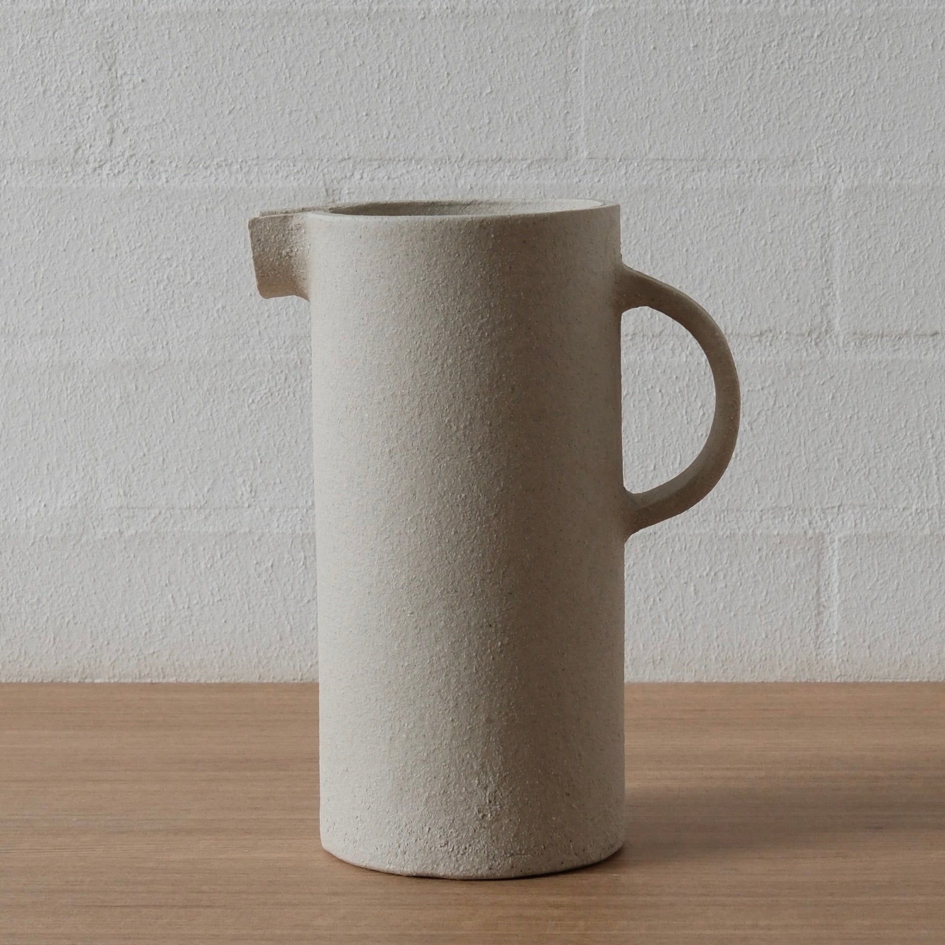 A white matte ceramic pitcher with a textured finish, featuring a small integrated spout and a loop handle, standing on a light wood surface against a white brick wall.