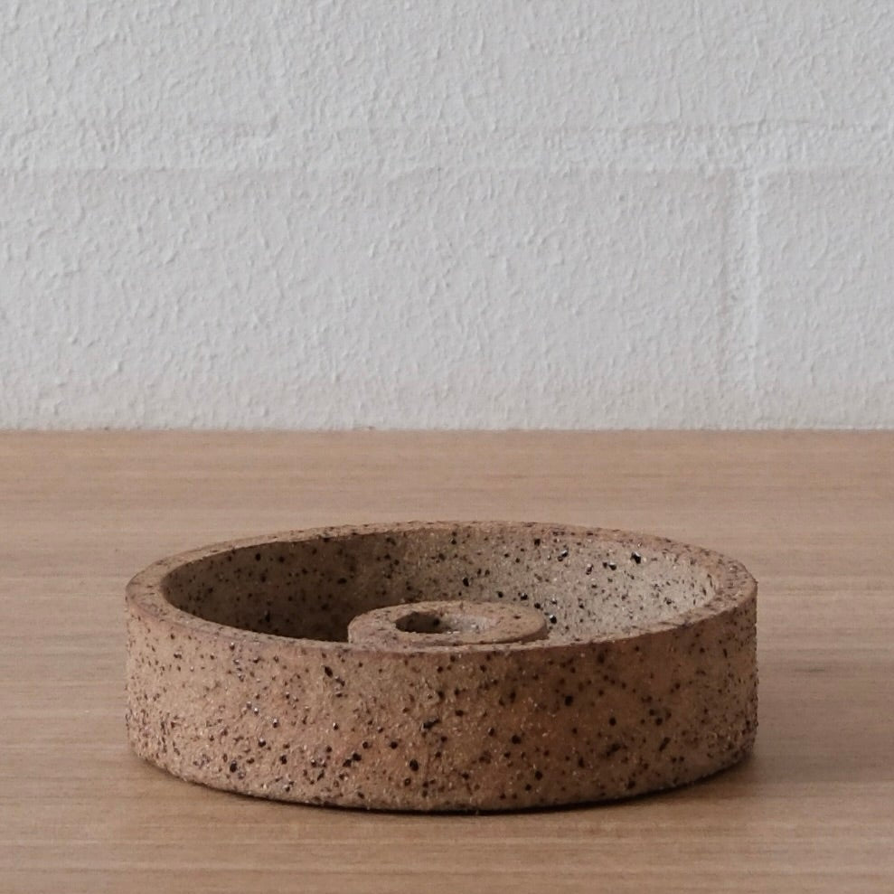 A round ceramic candle holder with a brown speckled finish, sitting on a light wood surface against a white brick wall background.