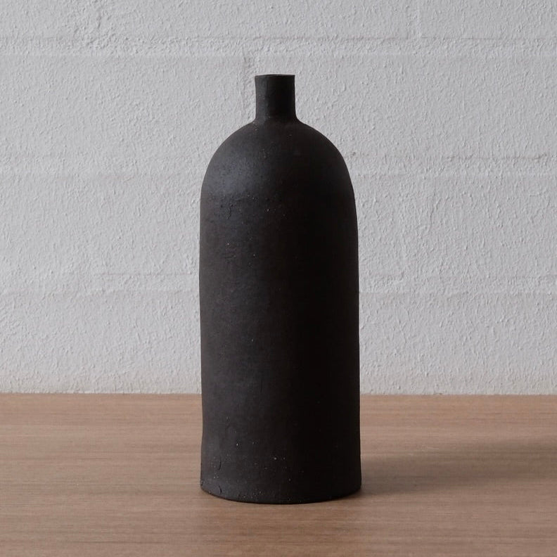 A matte black ceramic bottle vase with a rounded body and short neck, standing on a light wood surface against a white brick wall.