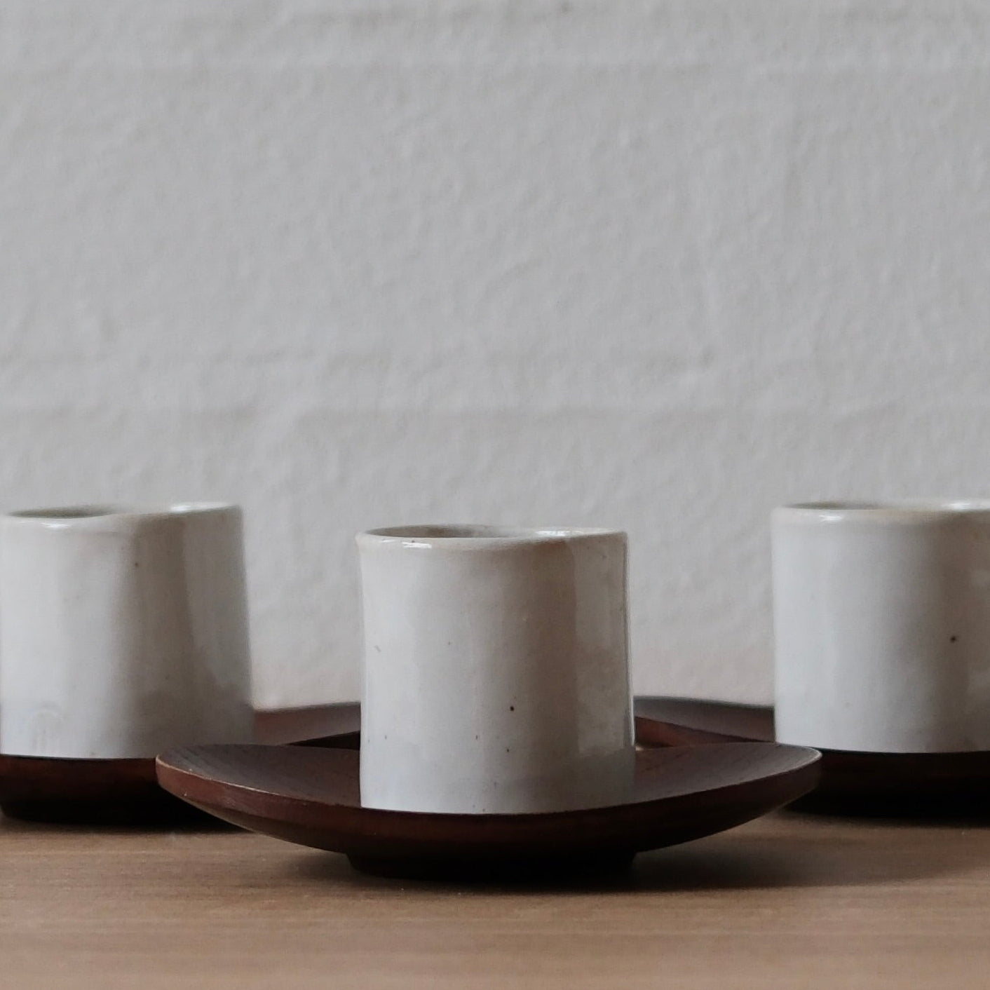 Three white ceramic espresso cups on dark wooden saucers against a white textured wall.